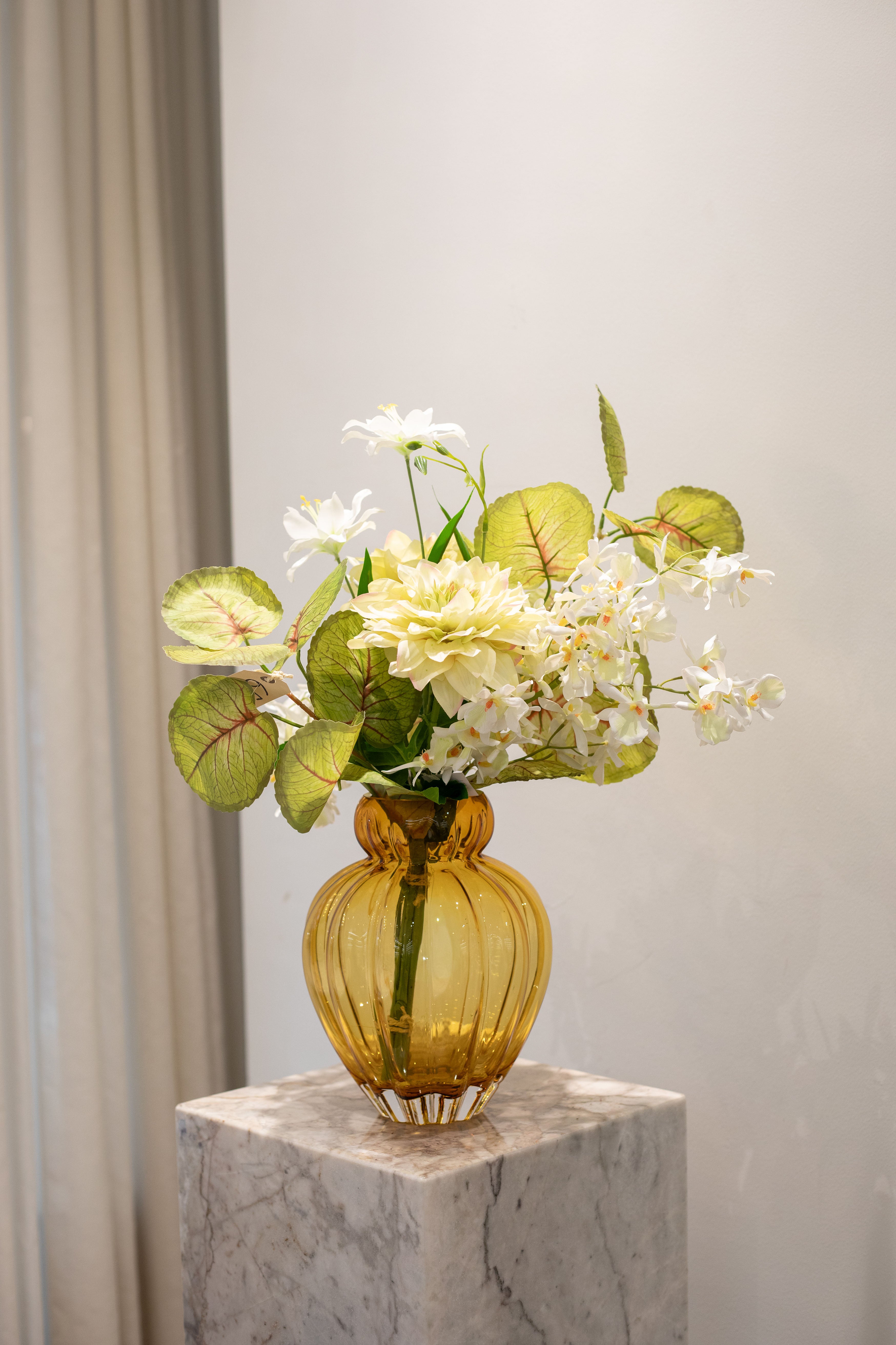 Billede af kunstig buket i hvide og grønne nuancer, der er placeret i en orange glas vase, der står ovenpå en marmor pedestal.