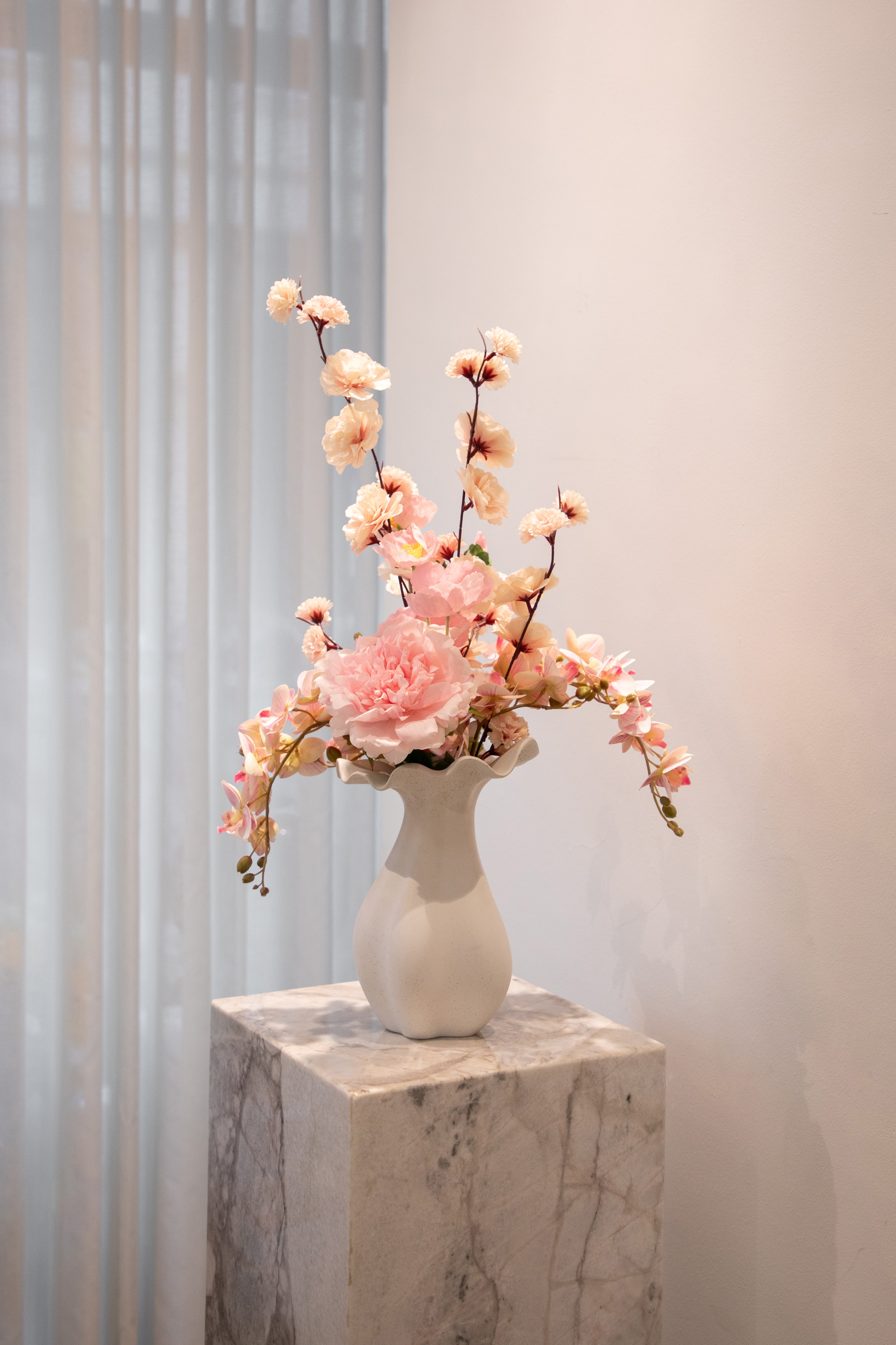 Kunstig buket i lyserøde nuancer, placeret i en hvid keramik vase, der står på en marmor pedestal.