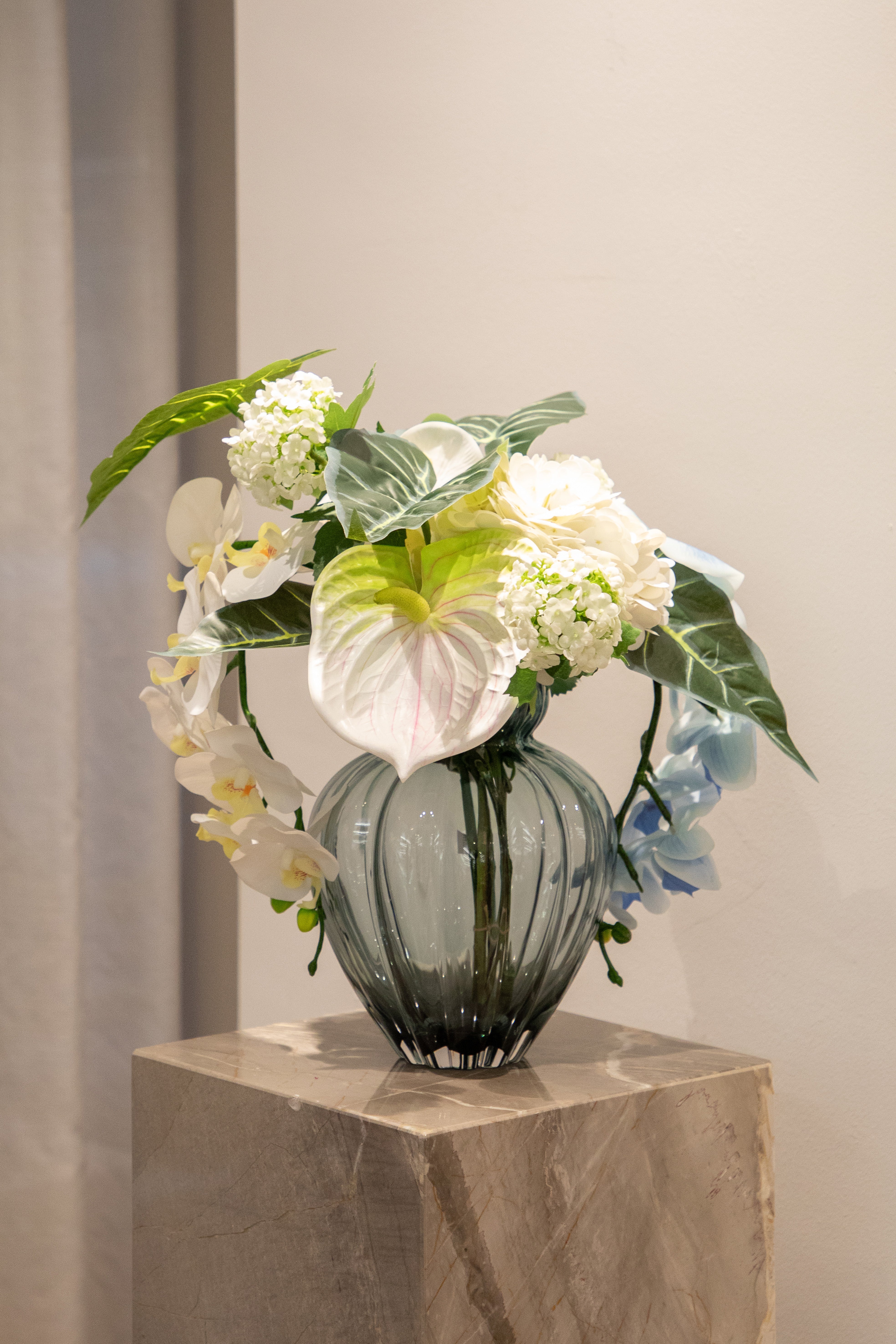 Kunstig buket i hvide, grønne og blå nuancer, der står i en blå glas vase, ovenpå en marmor piedestal.