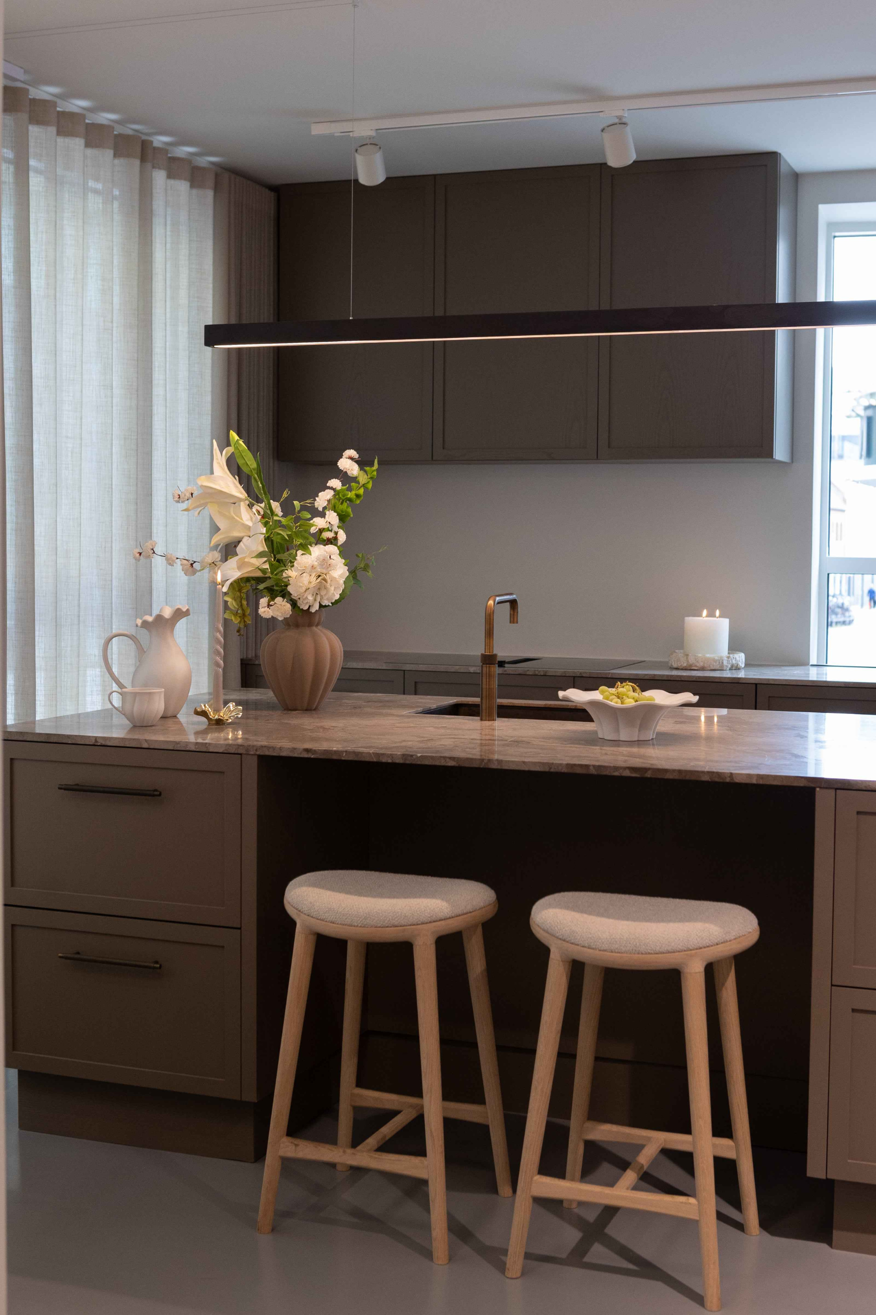 Moderne køkken med mørkebrune skabslåger og køkkenø i natursten. På øen står to lyse barstole med polstrede sæder, en vase med hvide blomster, en hvid kande og en dekorativ skål. Over øen hænger en lang, sort lampe, og i baggrunden ses et stort vindue med lyse gardiner samt en tændt hvid bloklys.