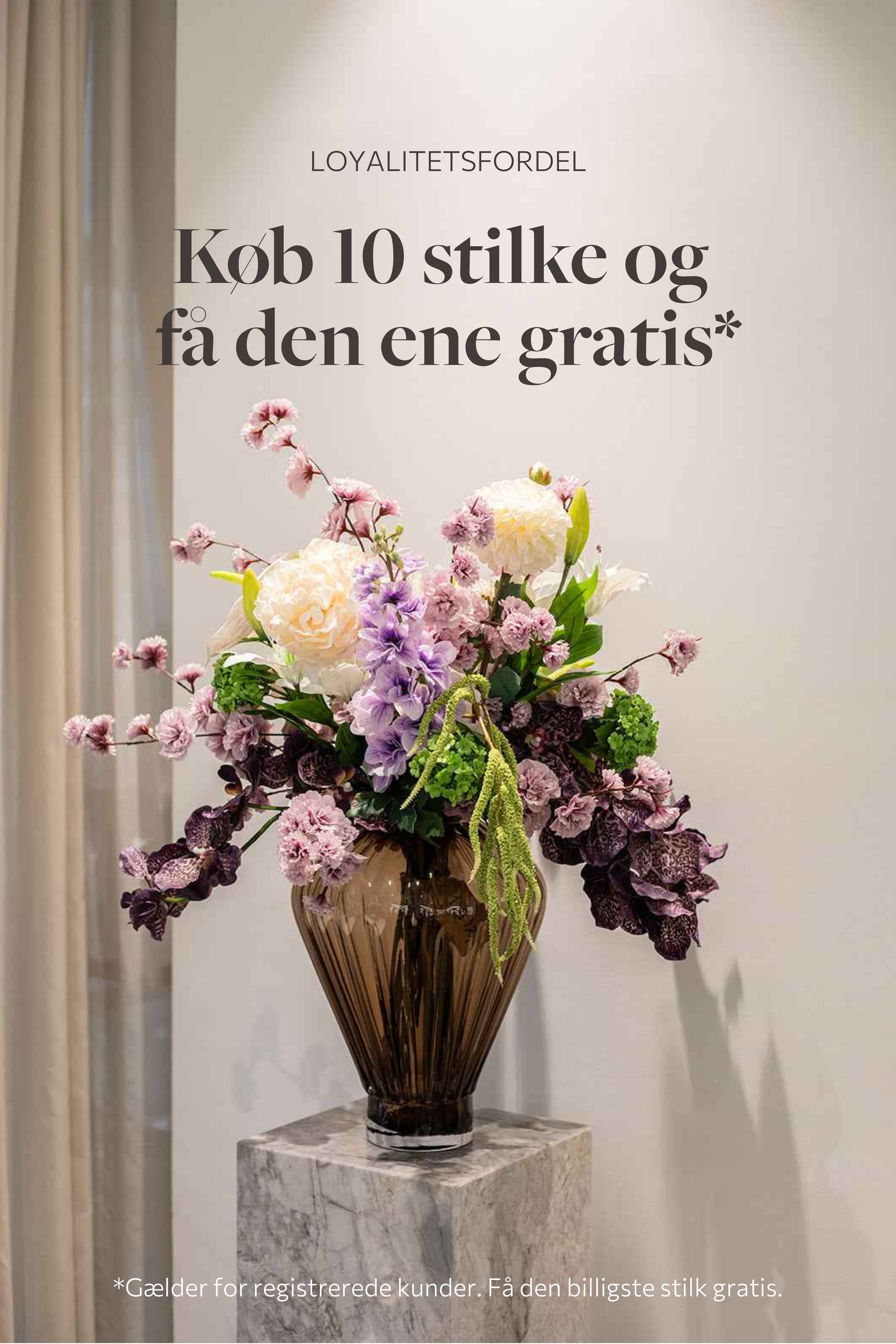 Stor farverig buket af kunstige blomster, placeret i en stor glasvase, der står på et marmor bord.