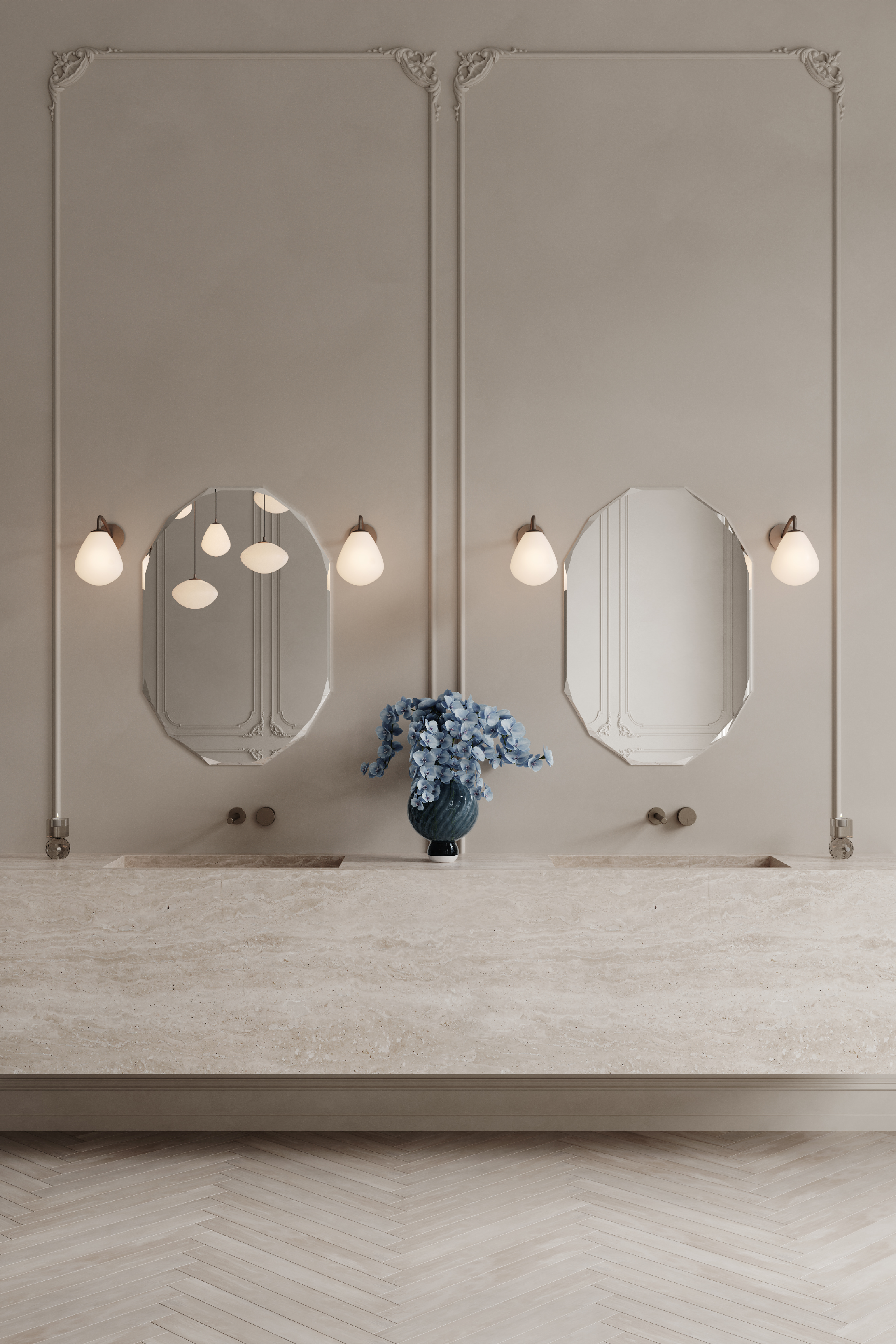 Badeværelse med lyse beige vægge og detaljerede paneler. To ovale spejle er placeret side om side, hver flankeret af to væglamper med hvide glasskærme. Under spejlene ses en stor bordplade i natursten med indbyggede vaske. På midten af bordpladen står en mørk vase med blå blomster, og gulvet er lagt i sildebensmønster.
