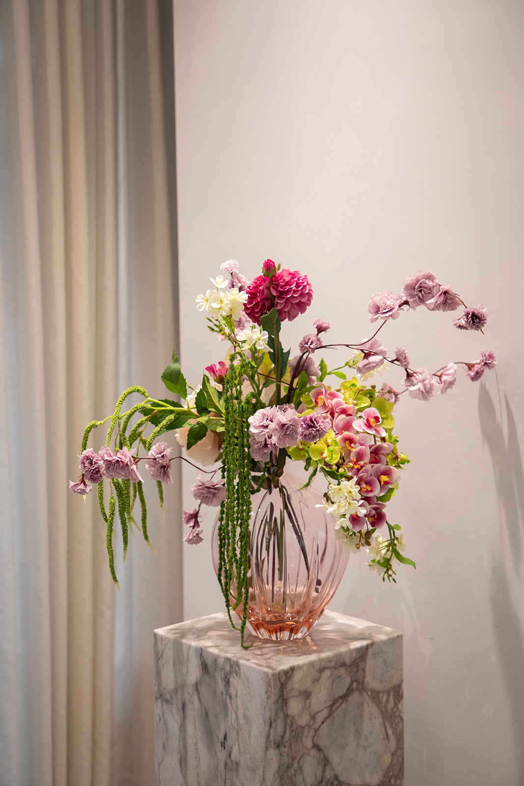 Buket af kunstige blomster i rosa, lilla og grønne nuancer. Placeret i en lyserød glasvase, der står på en marmor pedestal.