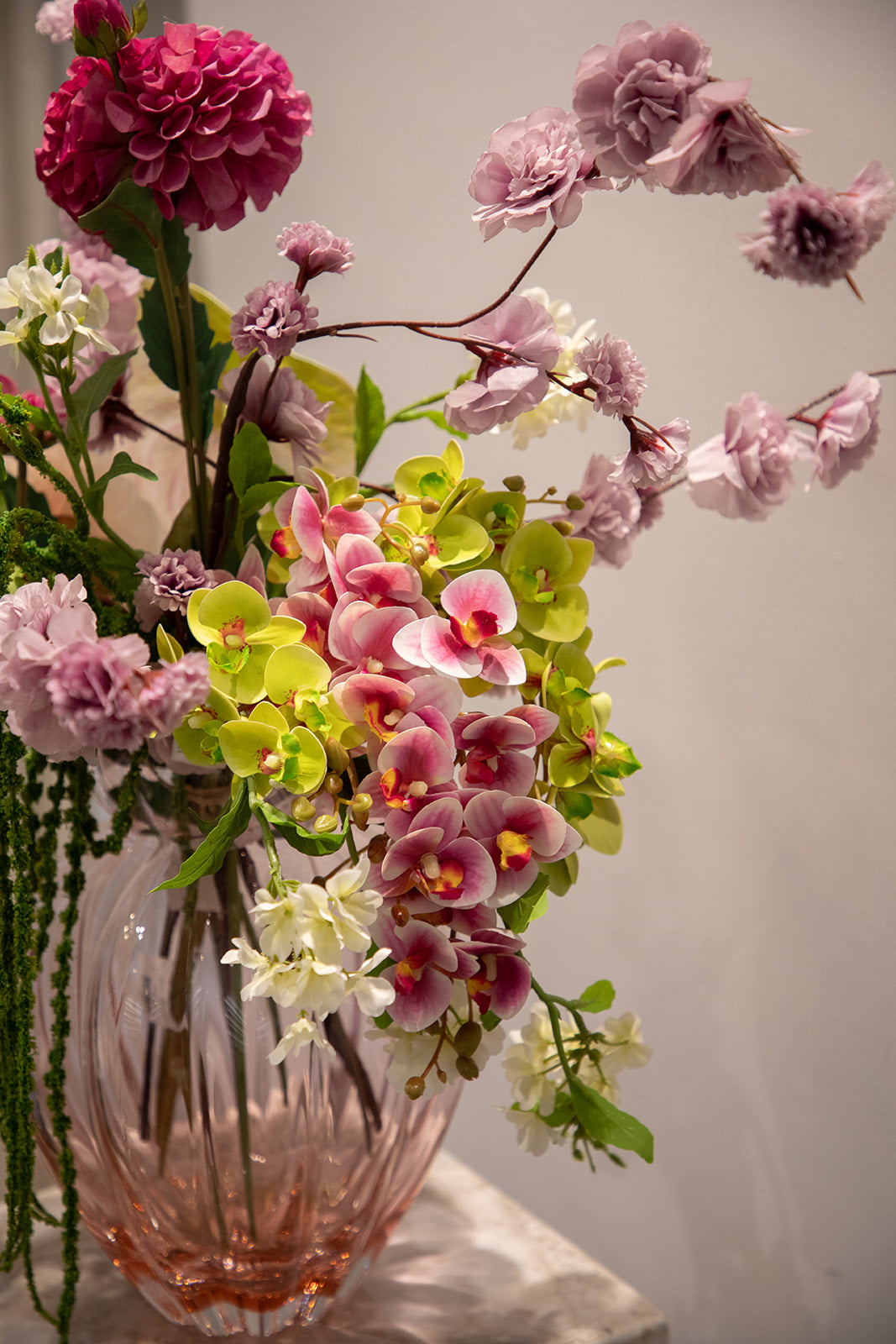 Nærbillede af buket af kunstige blomster i lilla, rosa og grønne nuancer. Placeret i en lyserød glasvase, der står på en marmor pedestal.
