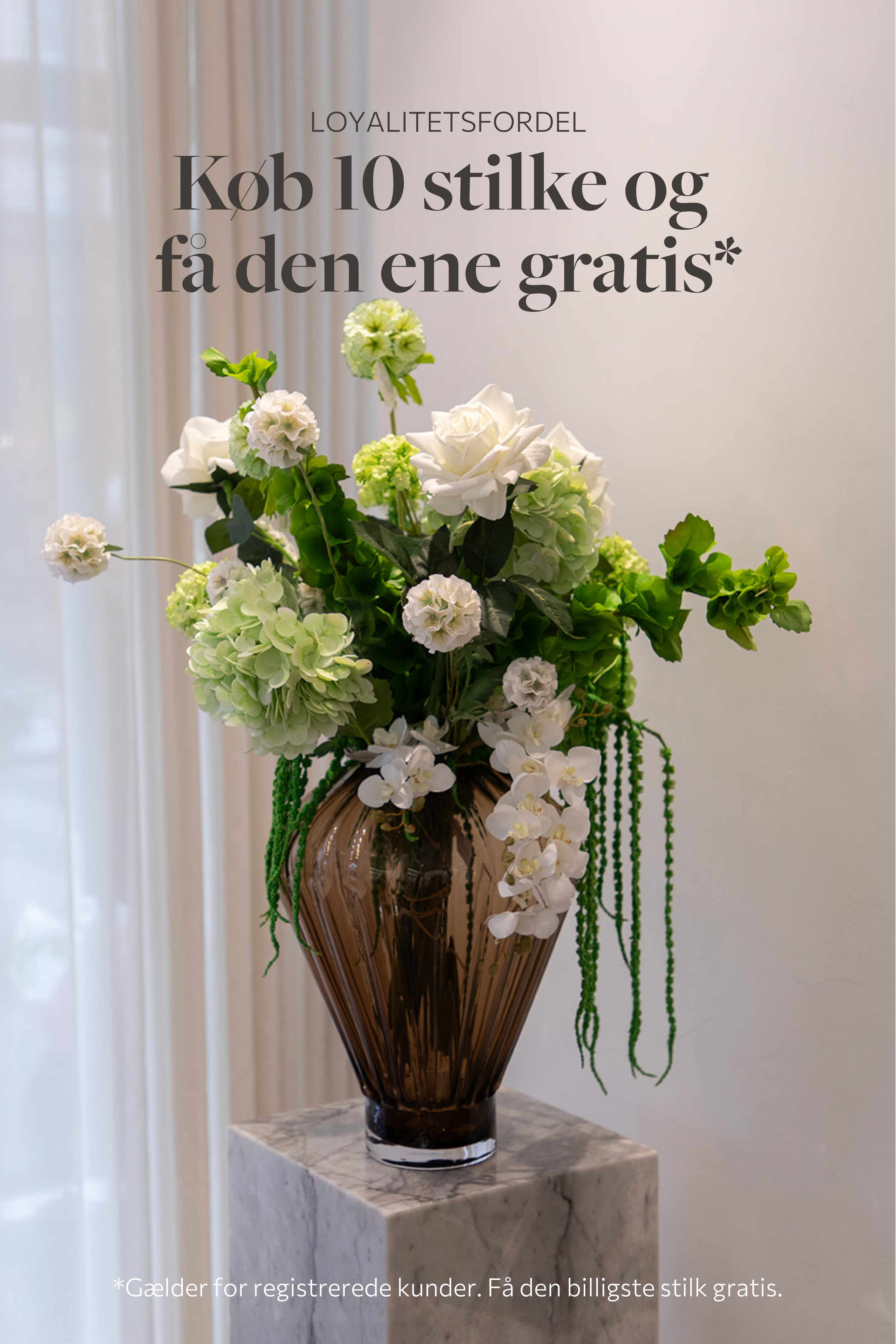 Stor buket af kunstige blomster i grønne og hvide nuancer. Placeret i en stor topaz farvet glasvase, står ovenpå en marmorpedestal