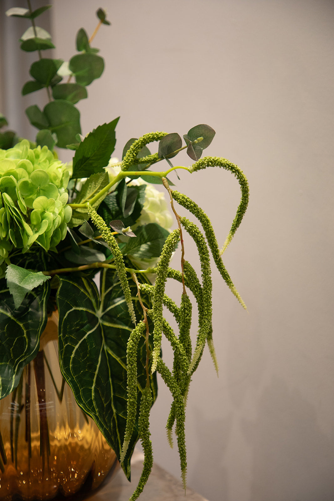 Nær billede af en kunstig buket i grønne nuancer. Står i en orange farvet glasvase, der står oven på en pedestal.