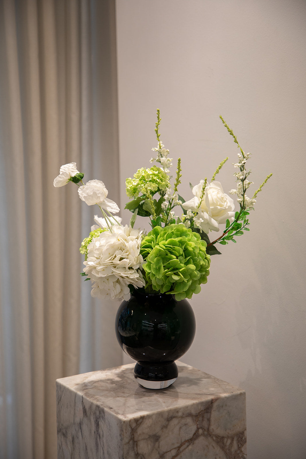 Buket af kunstige blomster i grønne og hvide nuancer. Placeret i en lille blå glasvase, der står på en marmor pedestal.
