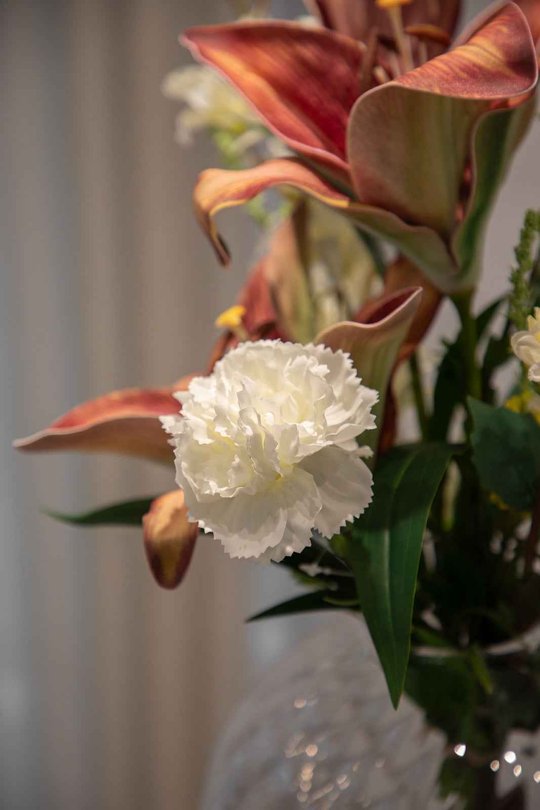 Nærbillede af buket af kunstige blomster i røde, cremfarvede og grønne nuancer, der er placeret i en gennemsigtig glasvase.