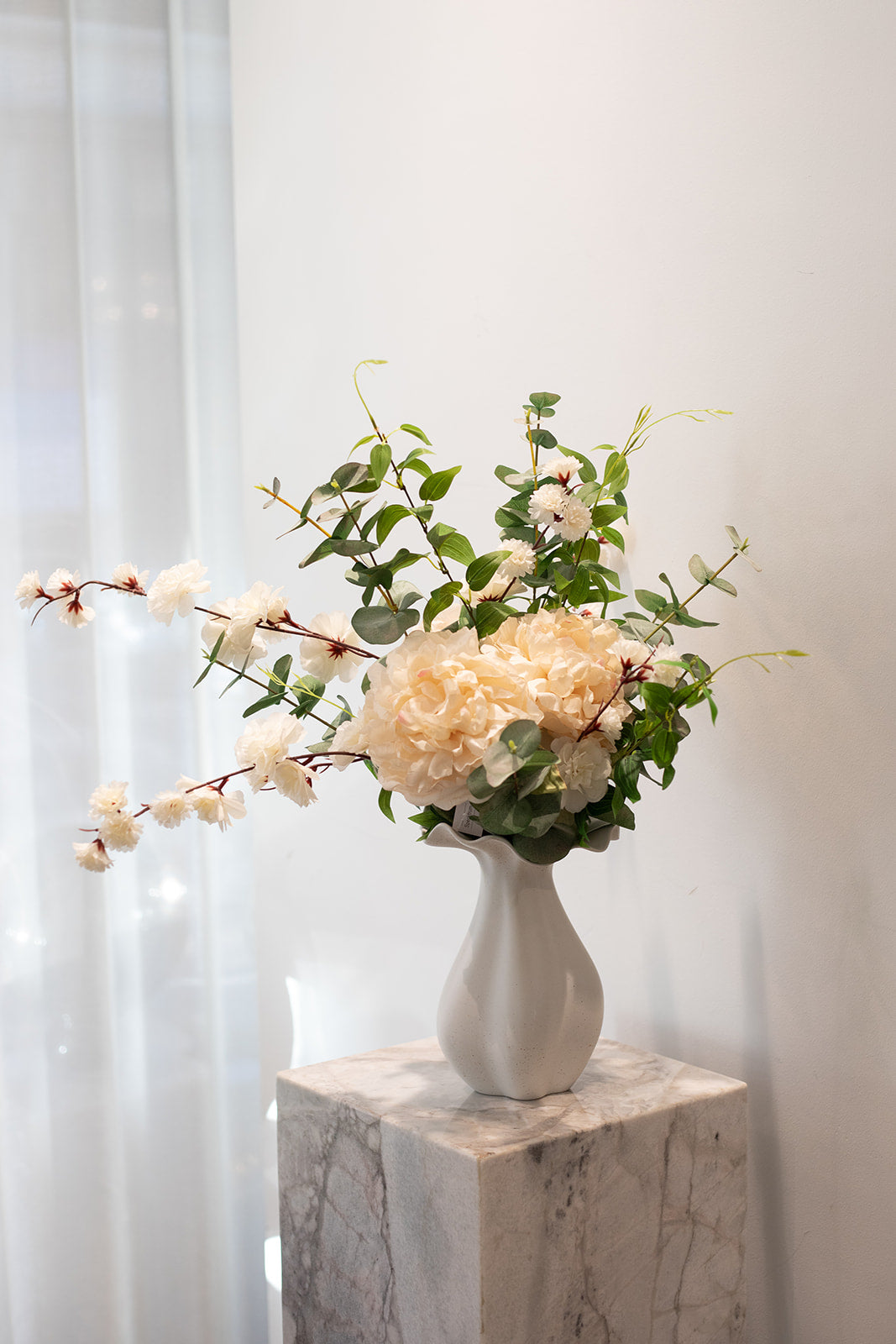 Stor kunstig buket i grønne og hvide nuancer, placeret i en hvid keramik vase, der står på en marmor pedestal.