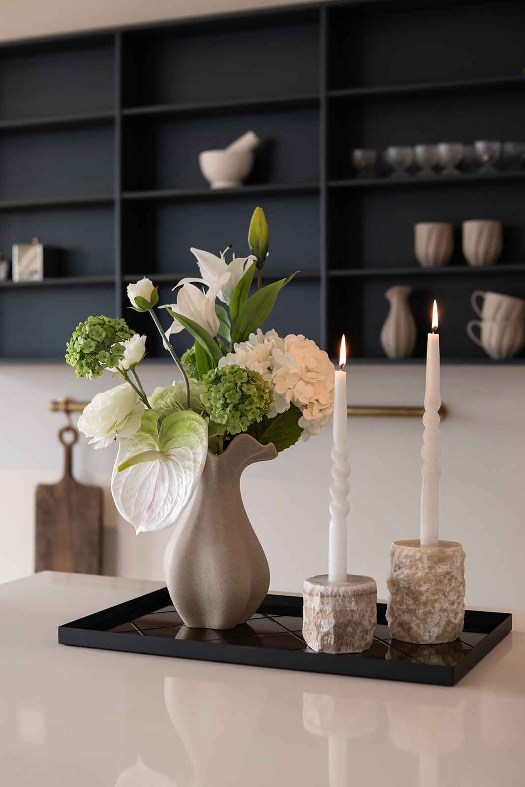 Beige vase med hvide liljer, hortensia og grønne blomster placeret på en sort spejlbakke. Ved siden af står to tændte hvide snoede lys i lyse stenhåndlavede lysestager.