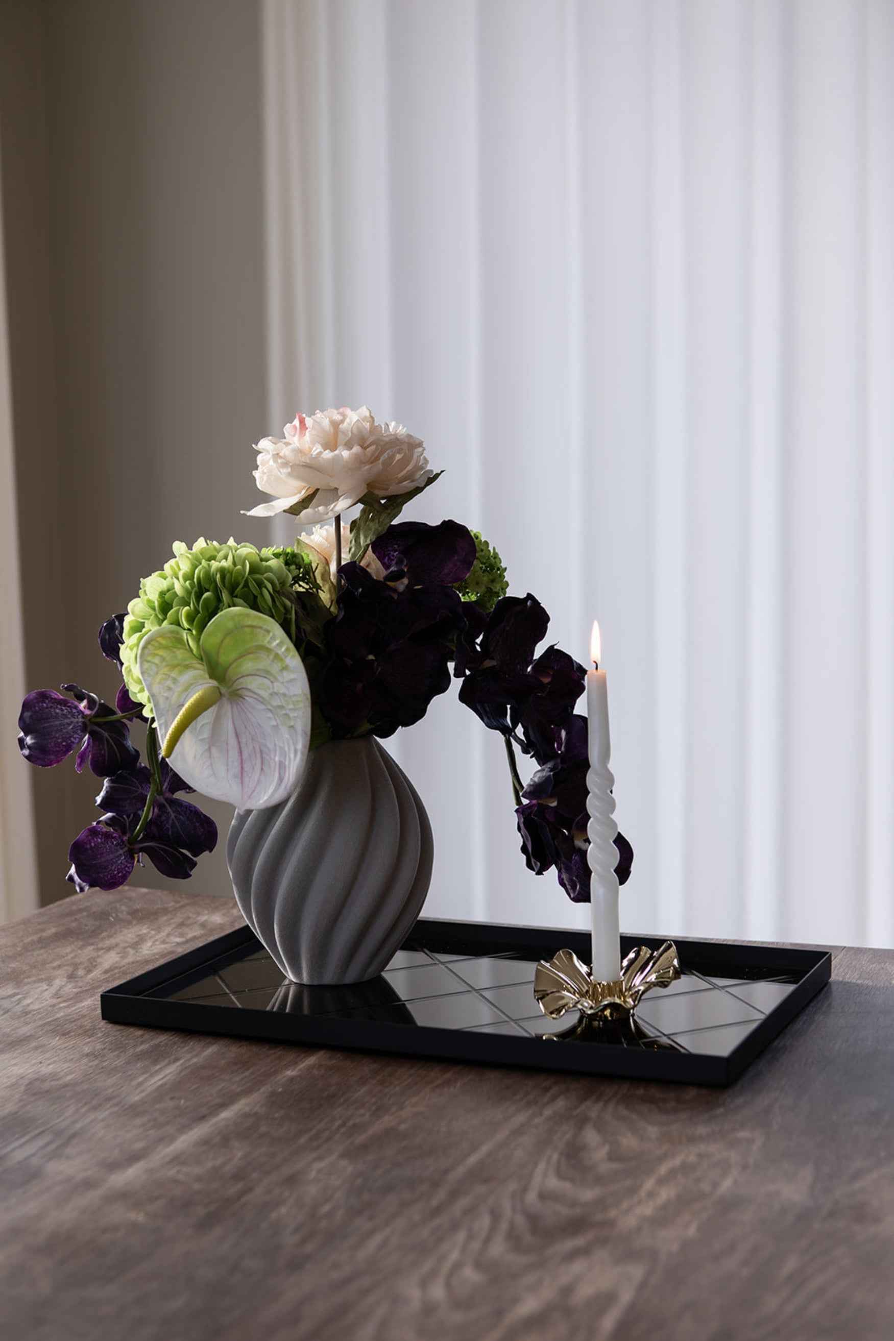 Gråriflet vase med blomsterarrangement bestående af lys rosa pæon, grønne blomster, hvid anthurium og mørkelilla orkidéer placeret på en sort spejlbakke. Ved siden af står et tændt snoet hvidt lys i en guldfarvet lysestage.