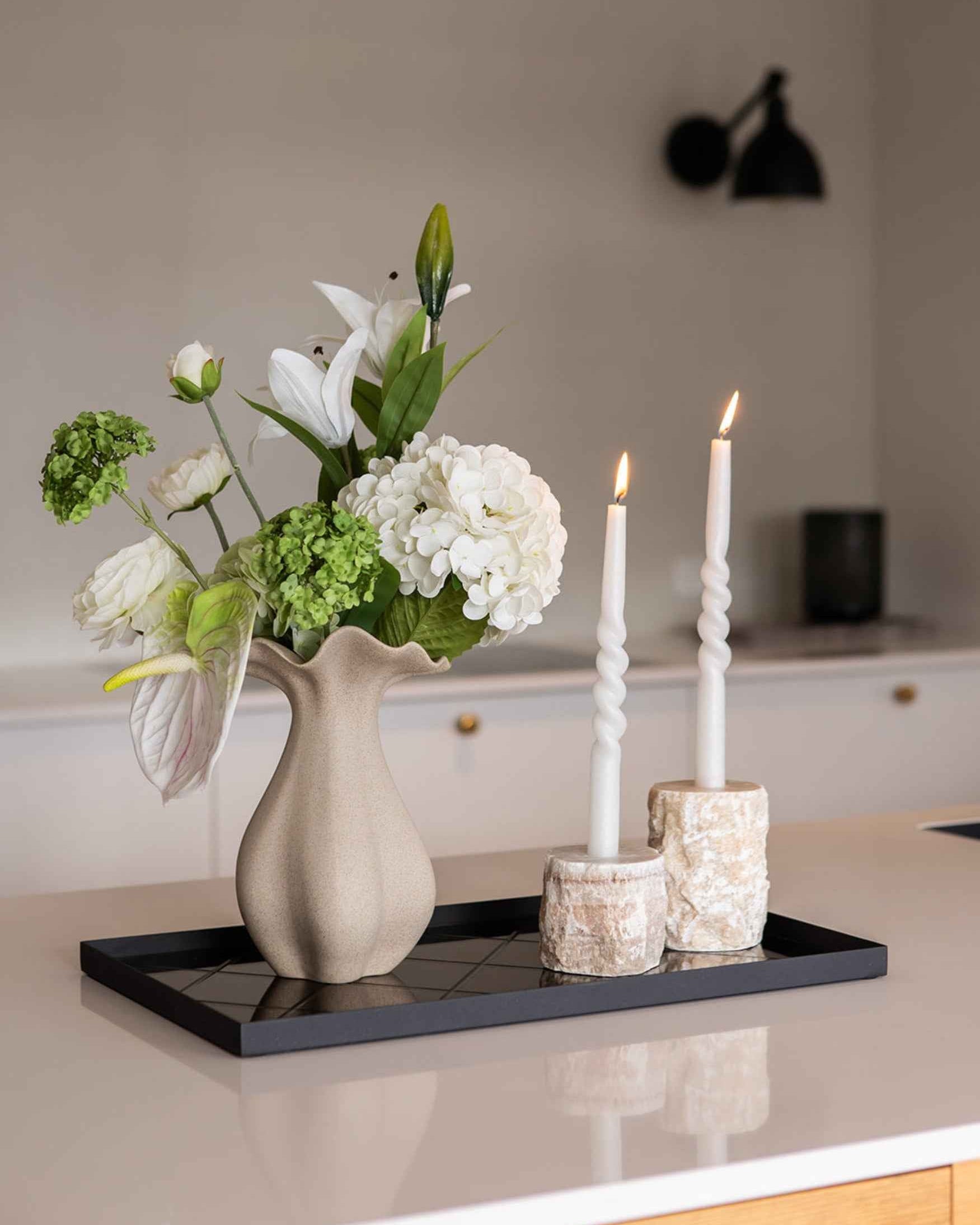 Beige vase med blomsterarrangement bestående af hvide liljer, hvide hortensia, hvide ranunkler, grønne blomster og en hvid anthurium placeret på en sort spejlbakke. Ved siden af står to tændte snoede hvide lys i lysestager af lys sten.