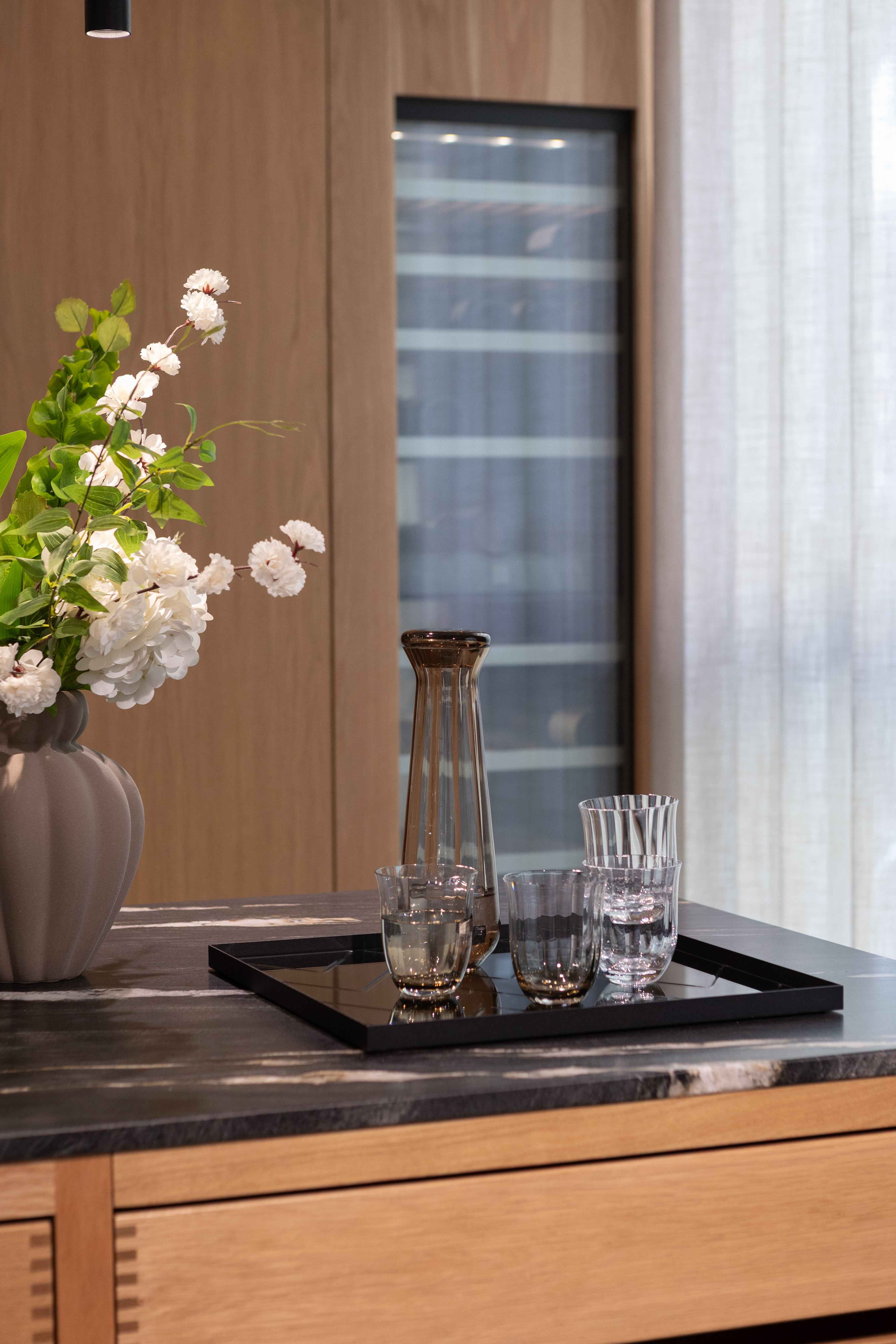 Rektangulær spejlbakke med sort kant placeret på en køkkenbordplade i mørk sten. På bakken står en høj, smal karaffel i røgfarvet glas og tre drikkeglas i forskellige former og mønstre.