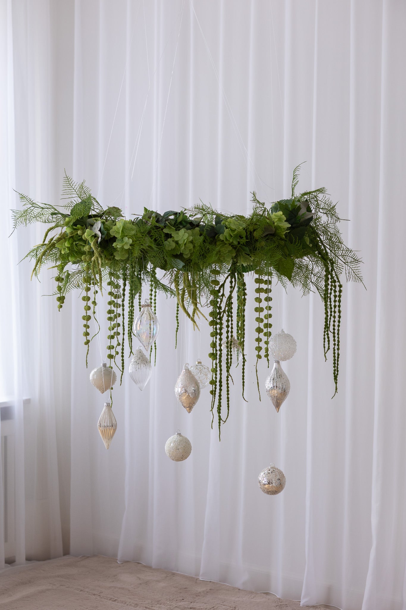 Grøn krans i kunstige blomster med hængende julekugler i hvide og sølv nuancer.