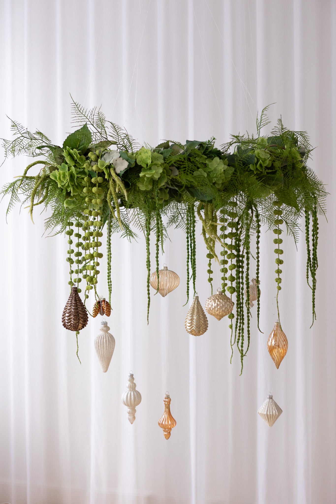 Stor hængende grøn krans af kunstige blomster med julekugler i guld, brune, og sølv nuancer.