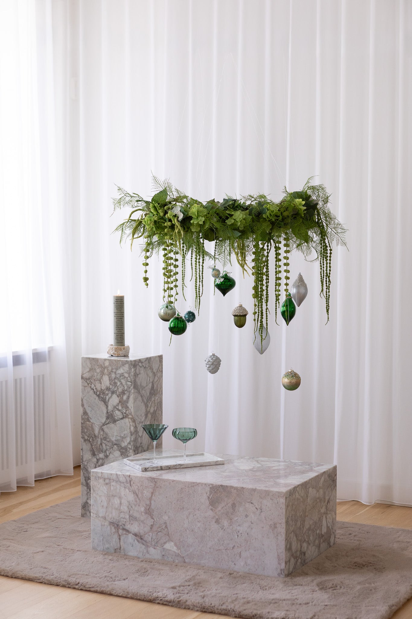 Stor hængende grøn krans af kunstige blomster med julekugler i grønne og sølv nuancer. Hænger over et marmorbord, ved siden af står en matchende pedestal med et kalenderlys.