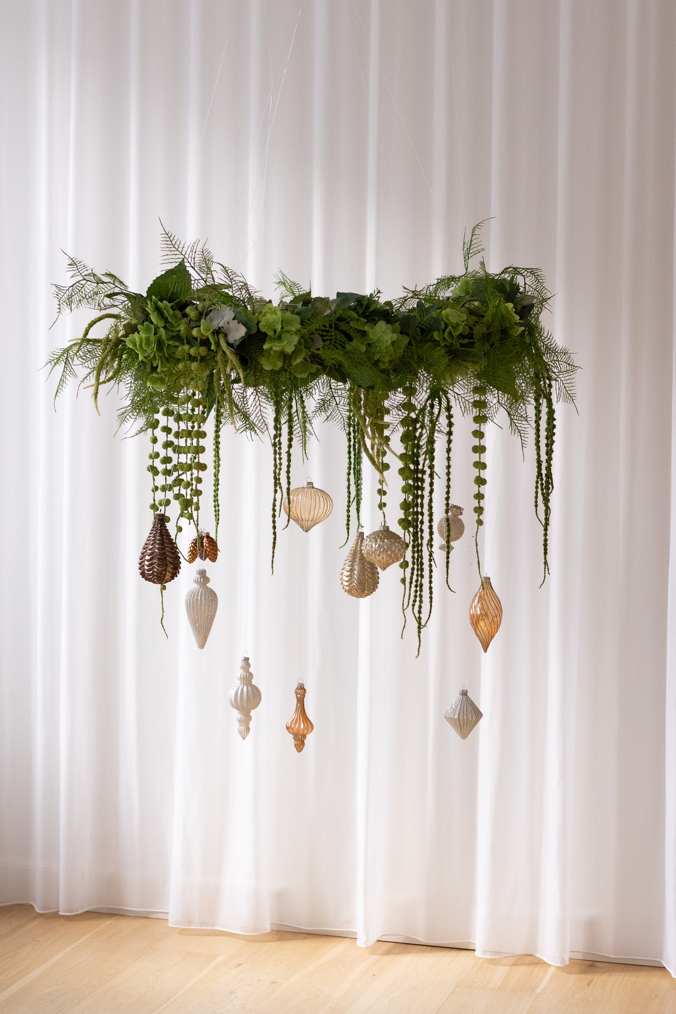 Stor hængende grøn krans af kunstige blomster med julekugler i guld, brune, og sølv nuancer.