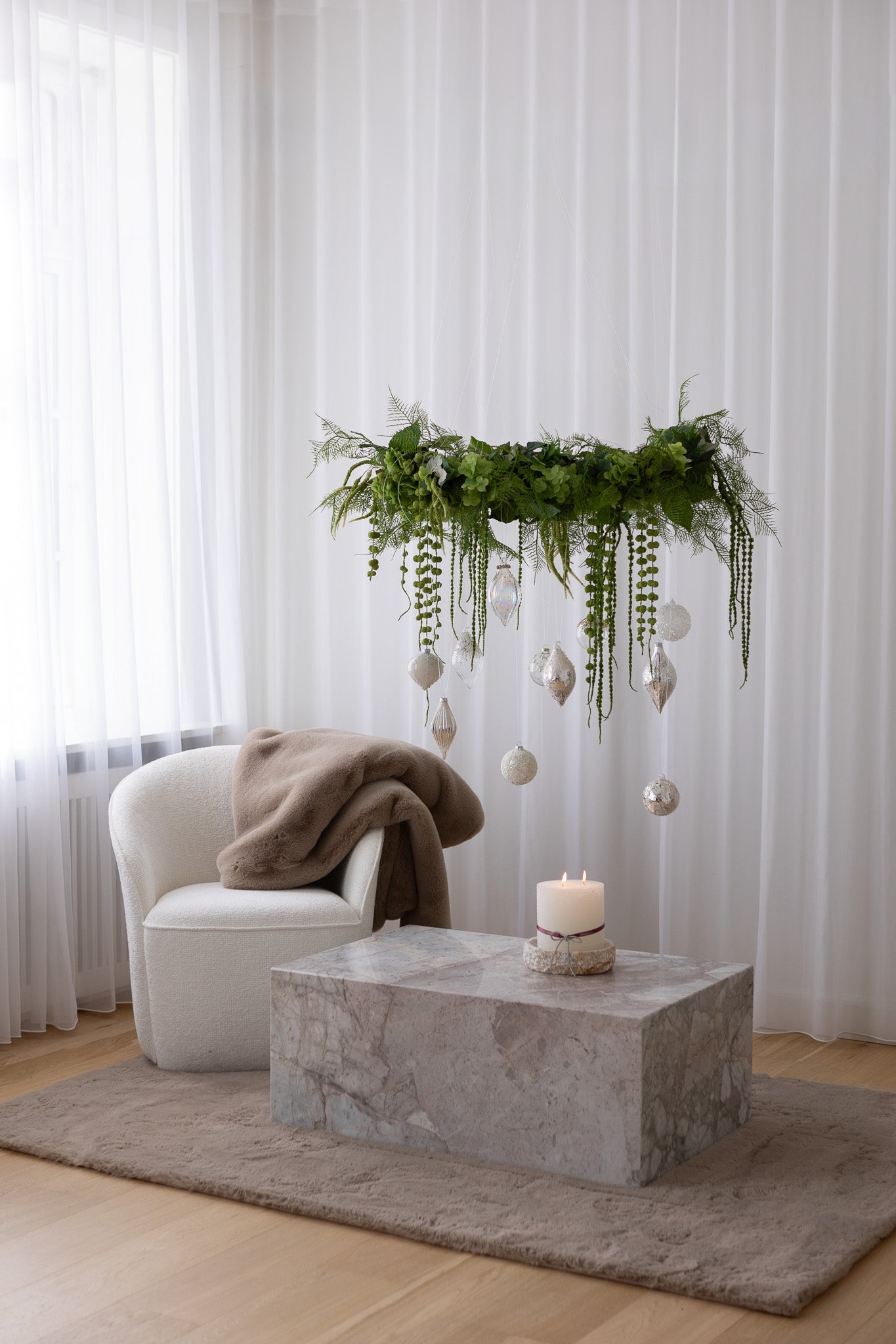 Krans i grønne blomster med hængende julekugler i hvide og sølv nuancer. Hænger over et marmorbord, ved siden af står en hvid loungestol.