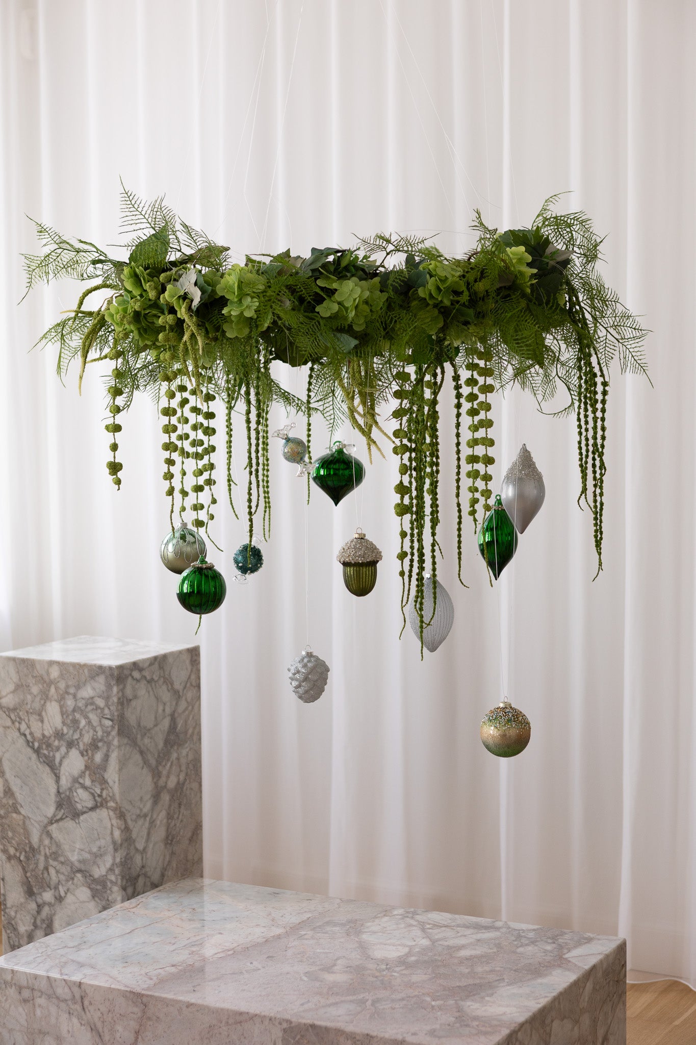 Stor grøn krans af kunstige blomster med grønne og sølv julekugler. Hænger over et marmorbord