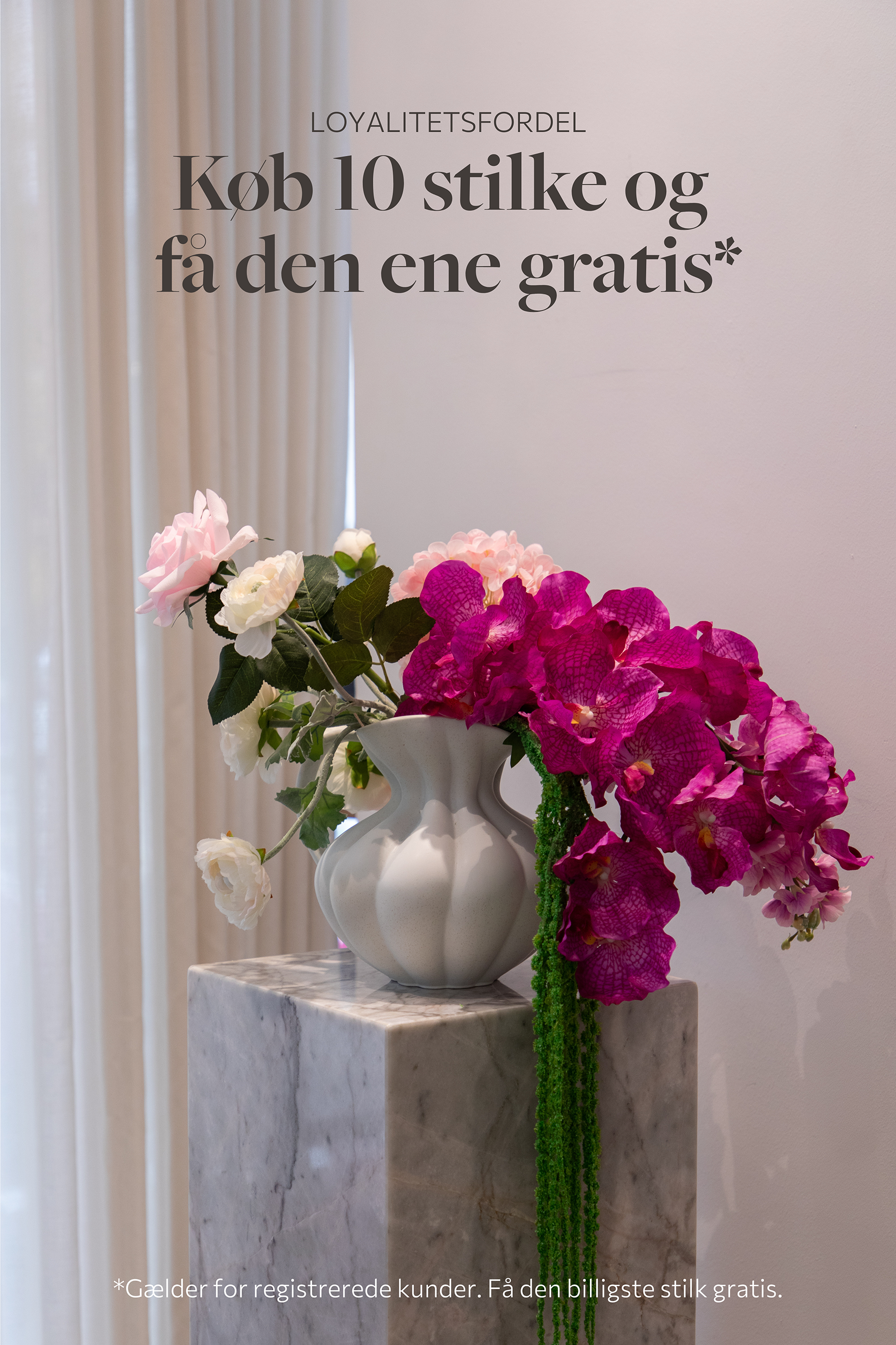 Stor farverig buket af kunstige blomster, placeret i en hvid keramik vase, der står på et marmorbord.