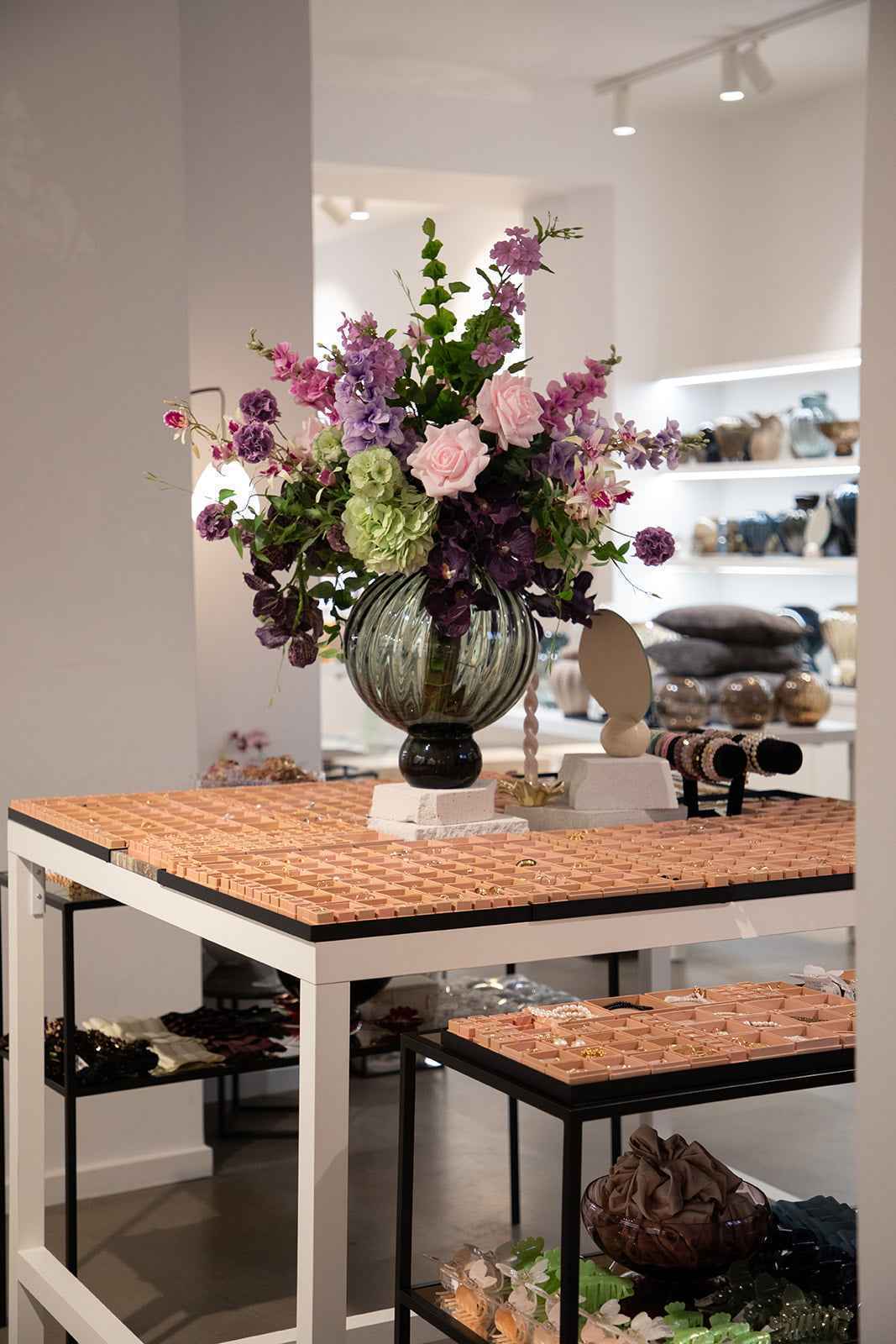 Et konsolbord med mange små smykkefag er dekoreret med en stor glasvase fyldt med farverige blomster. På og omkring bordet ses armbånd, hårpynt og andre accessories.