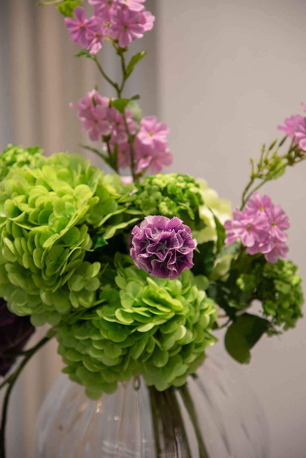 Nærbillede af buket af kunstige blomster i grønne og lilla nuancer, placeret i en gennemsigtige glasvase.