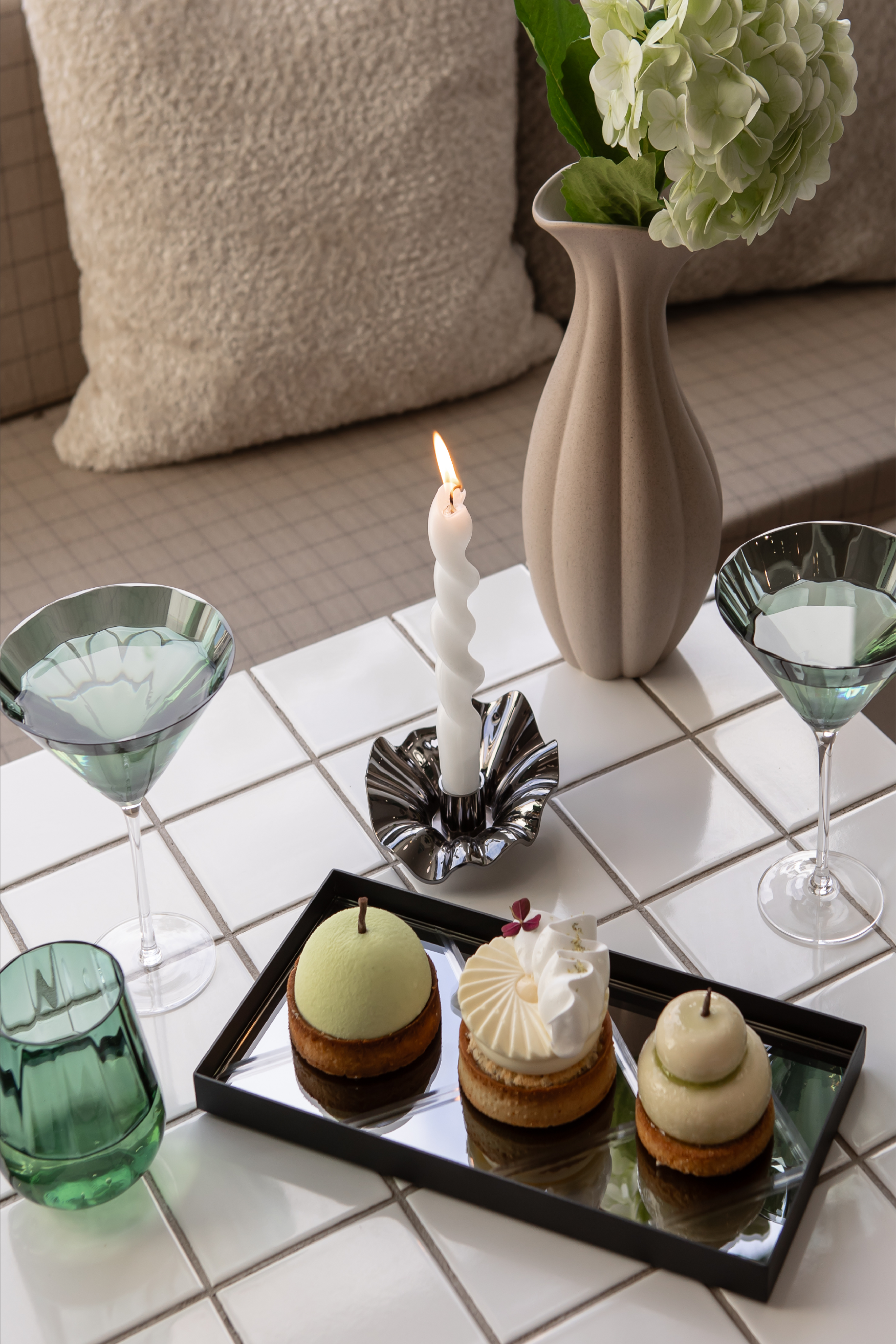 Et tændt hvidt snoet stearinlys står i en sort bølgeformet lysestage på et hvidt flisebord. Foran står et sort serveringsfad med tre små desserter, og omkring bordet er der grønne glas og grønne cocktailglas. Til højre ses en beige vase med en grøn hortensiablomst. I baggrunden er der puder på en sofa.