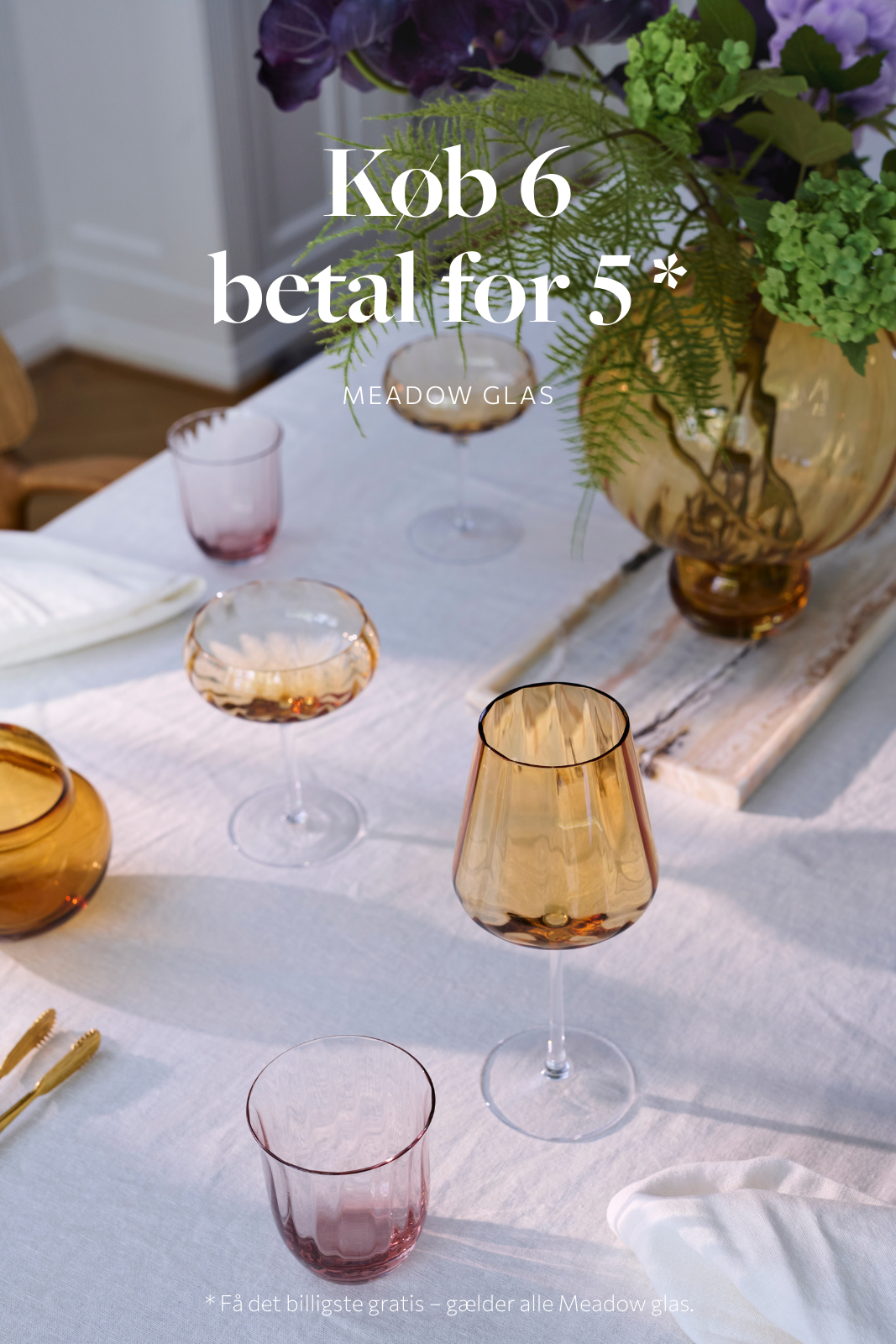 t smukt dækket bord med forskellige Meadow glas i gyldne og rosa nuancer. På billedet står både vinglas og cocktailglas fordelt på en hvid dug, sammen med servietter, guldbestik og en stor gul vase med grønne blade og lilla blomster. Teksten på billedet annoncerer en kampagne med ordene "Køb 6 betal for 5* Meadow glas", og nederst står der "*Få det billigste gratis – gælder alle Meadow glas".