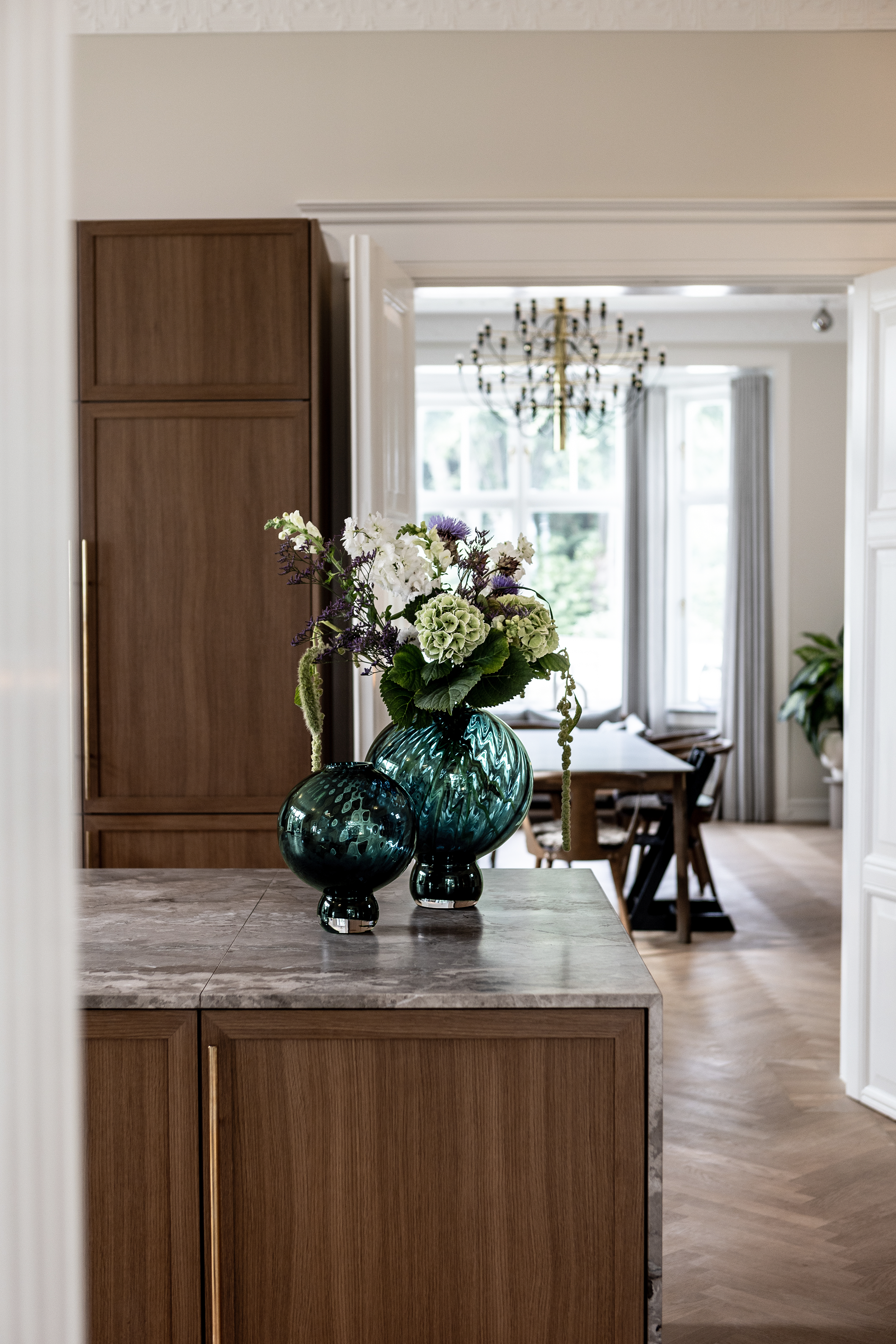 To grønne glasvaser med mønstret overflade står på en køkkenø i træ med grå marmorbordplade. Den største vase er fyldt med blomster og grønt løv. I baggrunden ses en spisestue med langbord, stole og en stor lysekrone samt et vindue med gardiner.