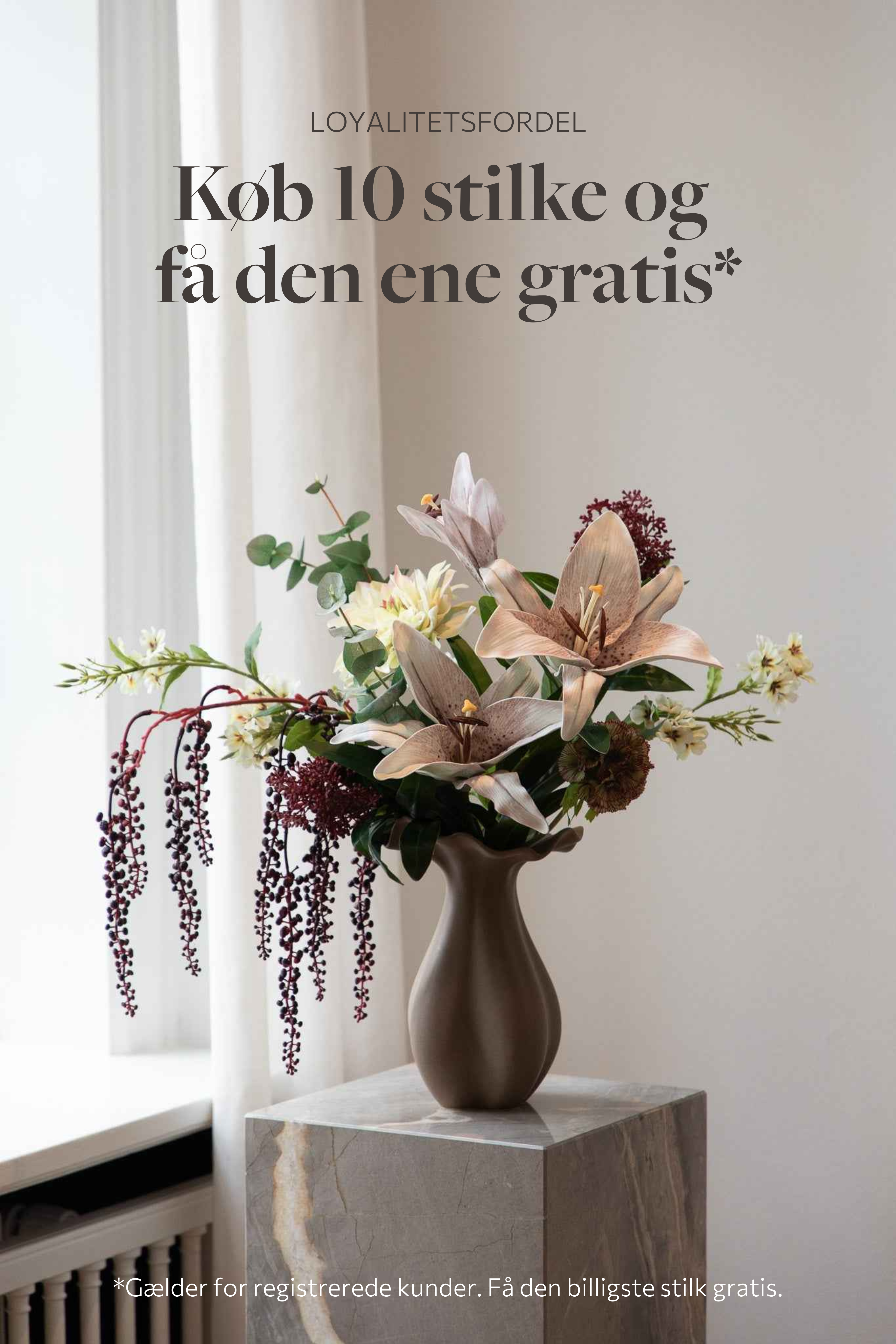 Farverig buket af kunstige blomster i røde, grønne og hvide nuancer. Placeret i en brun nellie keramikvase, står ovenpå en marmorpedestal.