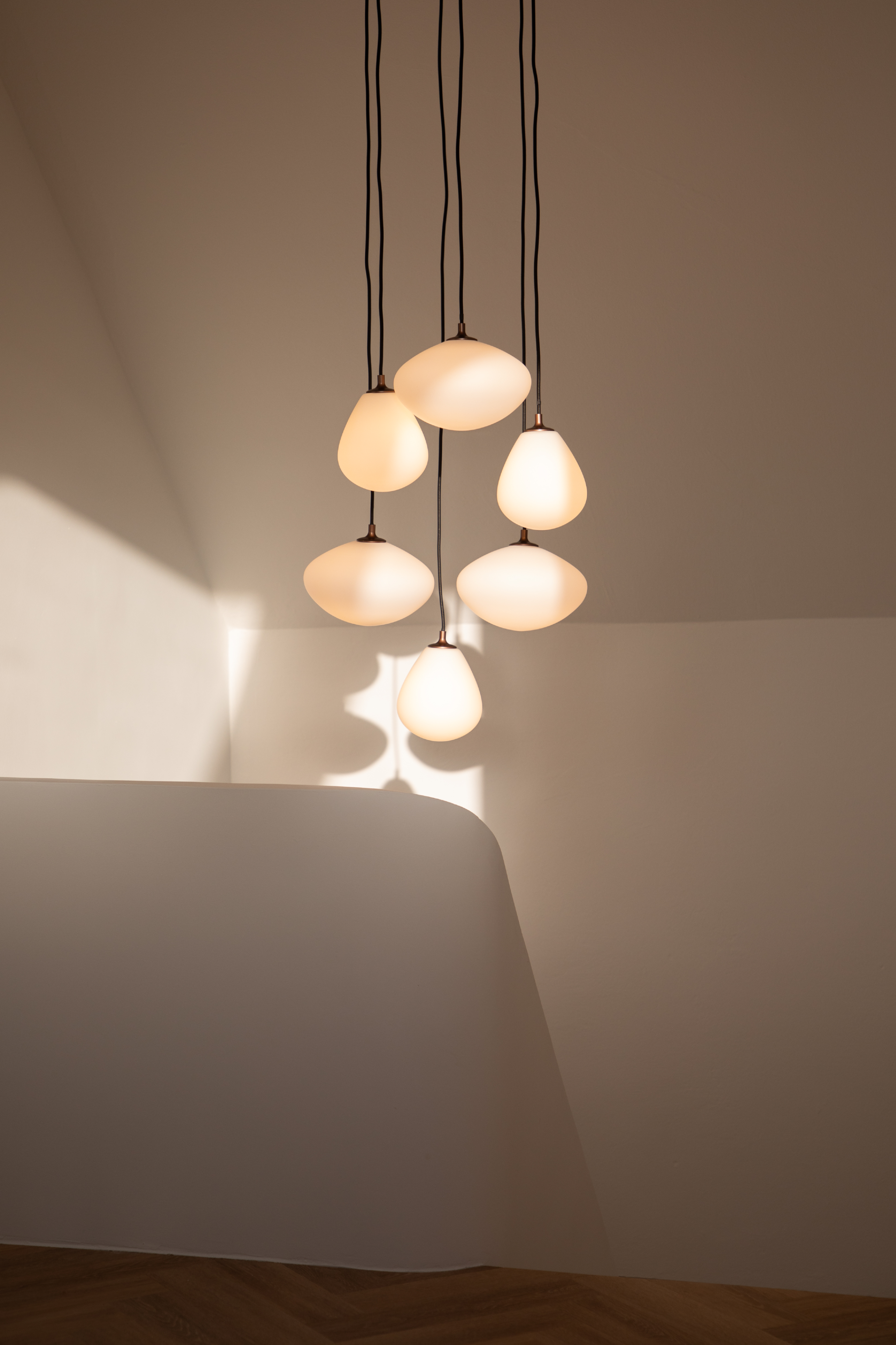 En klynge af seks pendellamper hænger fra loftet i sorte ledninger. Lamperne har dråbeformede og ovale hvide glasskærme, der spreder et varmt lys. De kaster bløde skygger på de lyse vægge og loftet, og under ses et trægulv i sildebensmønster.