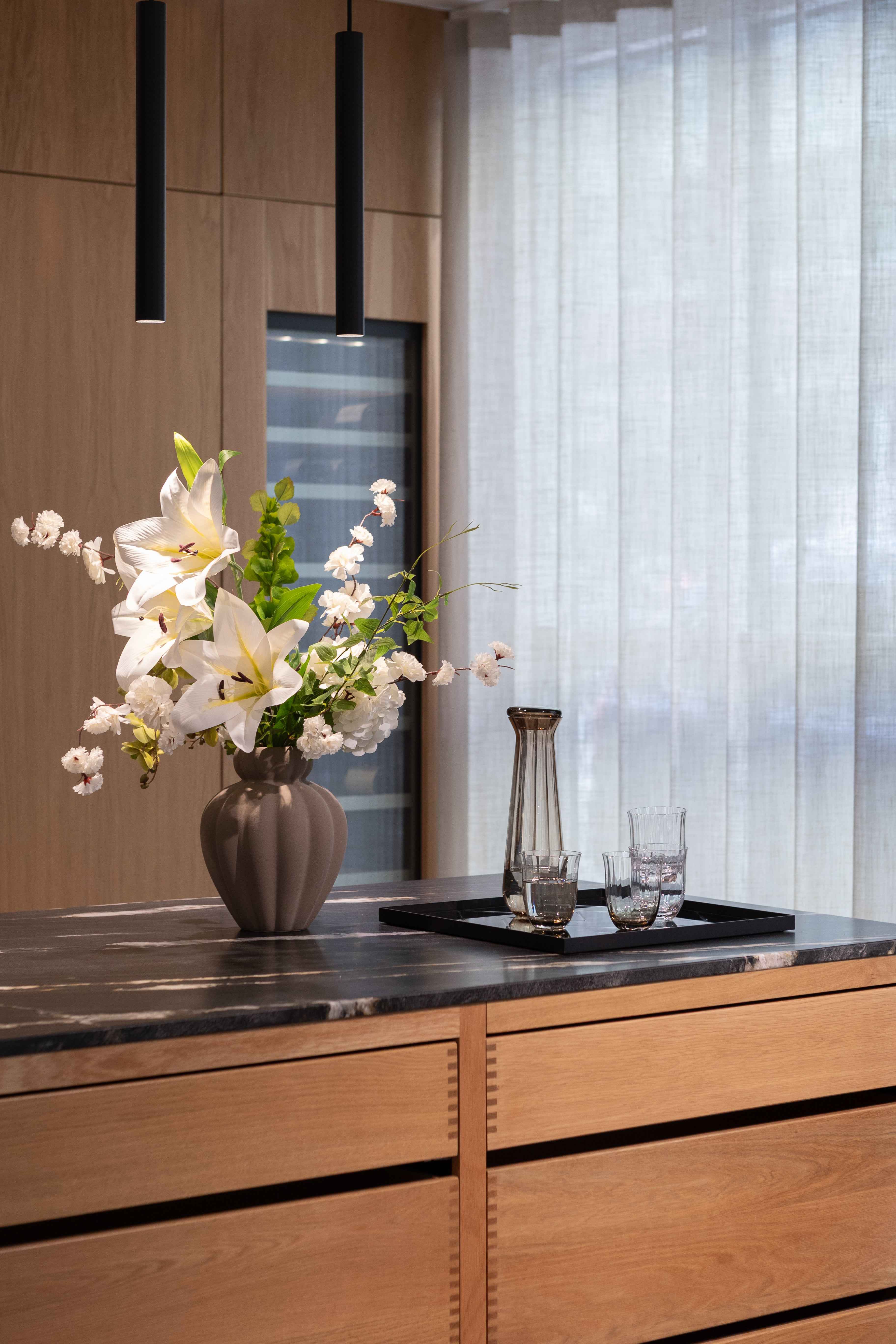 Rektangulær spejlbakke med sort kant placeret på en mørk køkkenbordplade. På bakken står en høj, smal karaffel i røgfarvet glas og to drikkeglas. Til venstre for bakken står en stor vase i grå keramik med hvide liljer og andre hvide blomster.