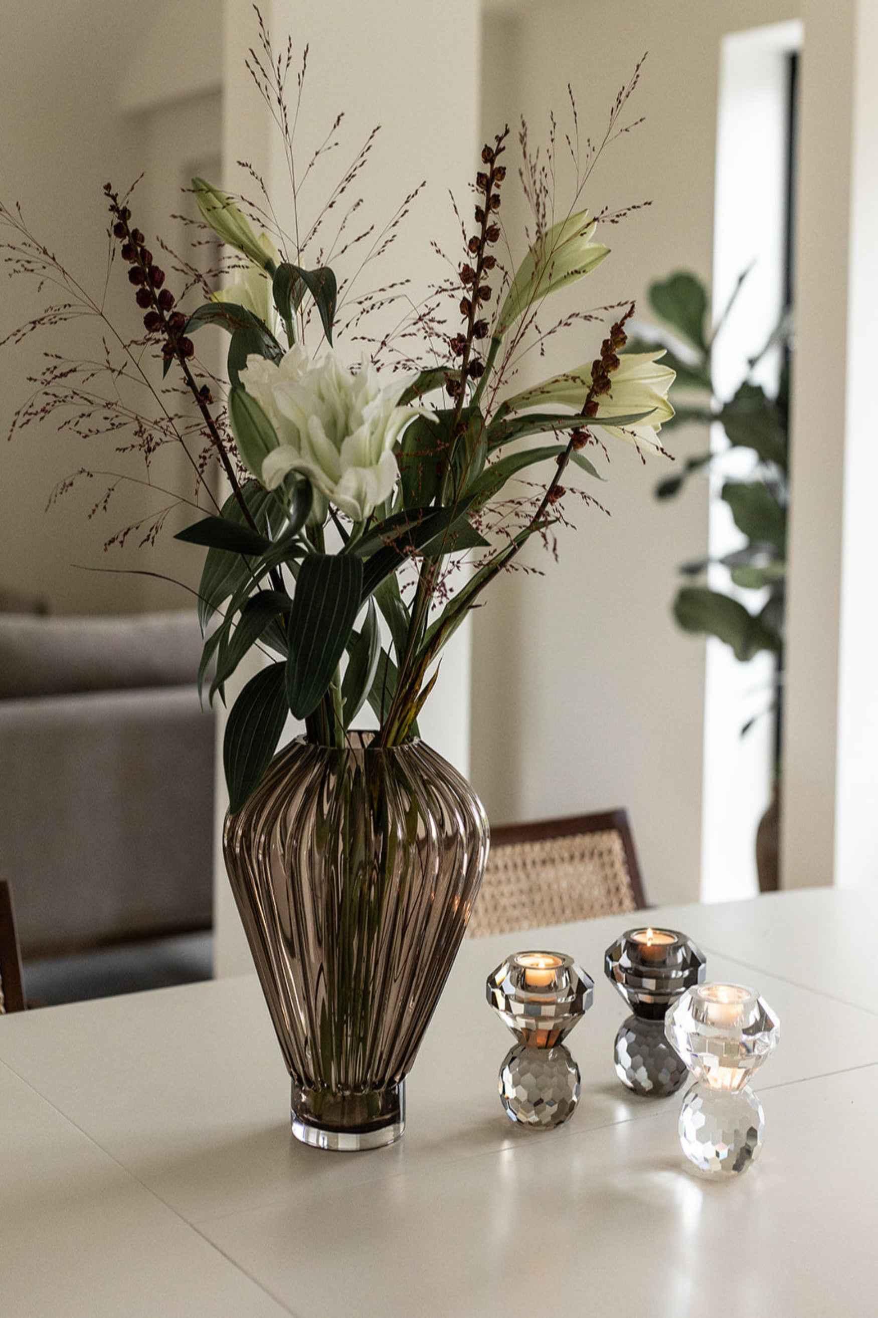 Et bord med en brun glasvase med hvide blomster og grønne blade. Ved siden af står tre fyrfadsstager i facetslebet krystal i sølv- og gråtoner, hver med et tændt fyrfadslys.