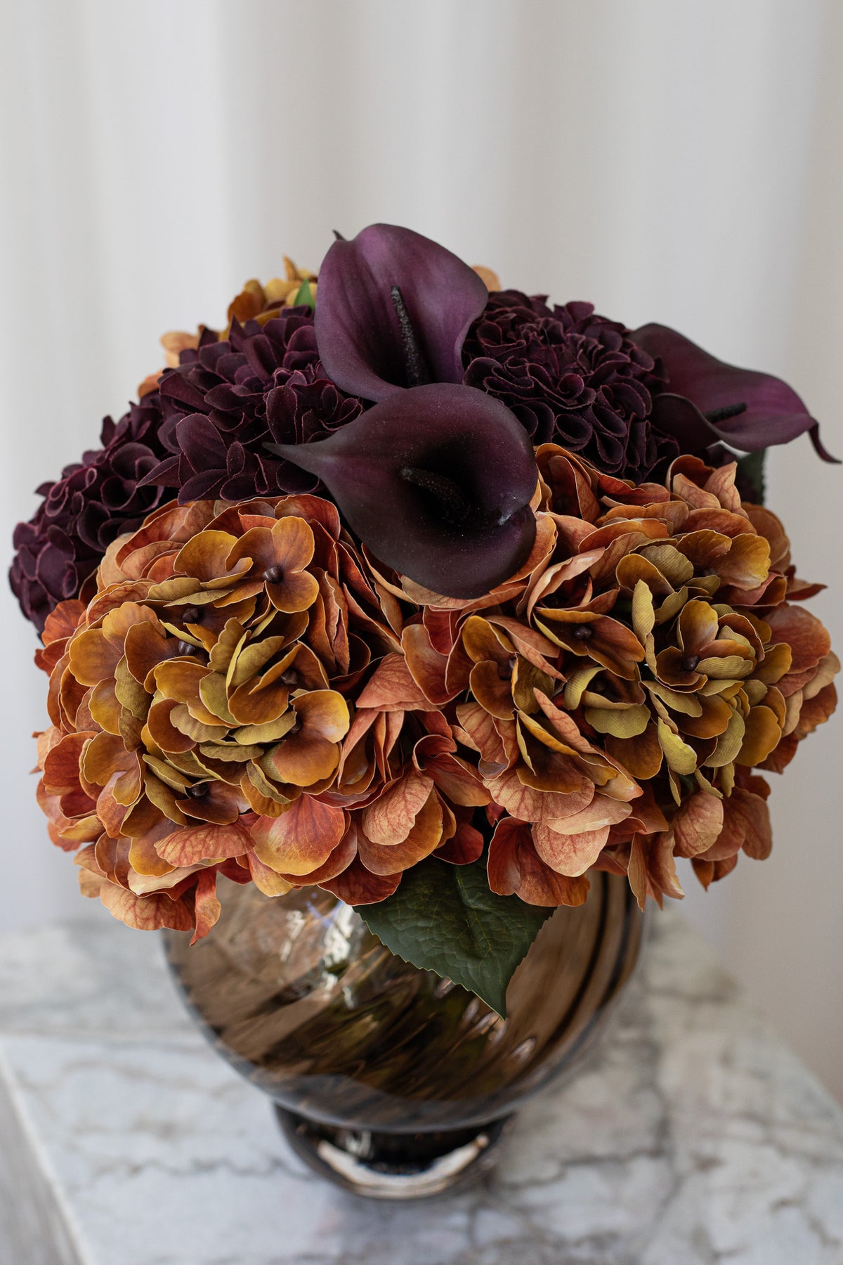 Kunstig buket med hortensia og calla liljer i brændt orange og lilla stylet i en brun glasvase.