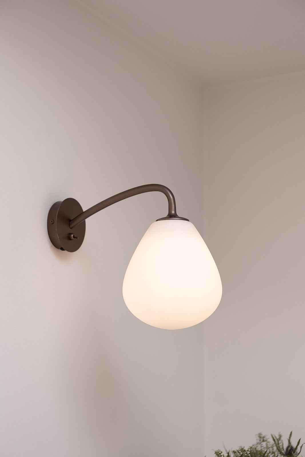 Nærbillede af en væglampe med rund vægplade i mørkt metal og en buet arm, der holder en dråbeformet hvid glasskærm. Lampen er monteret på en lys væg og giver et blødt, varmt lys.
