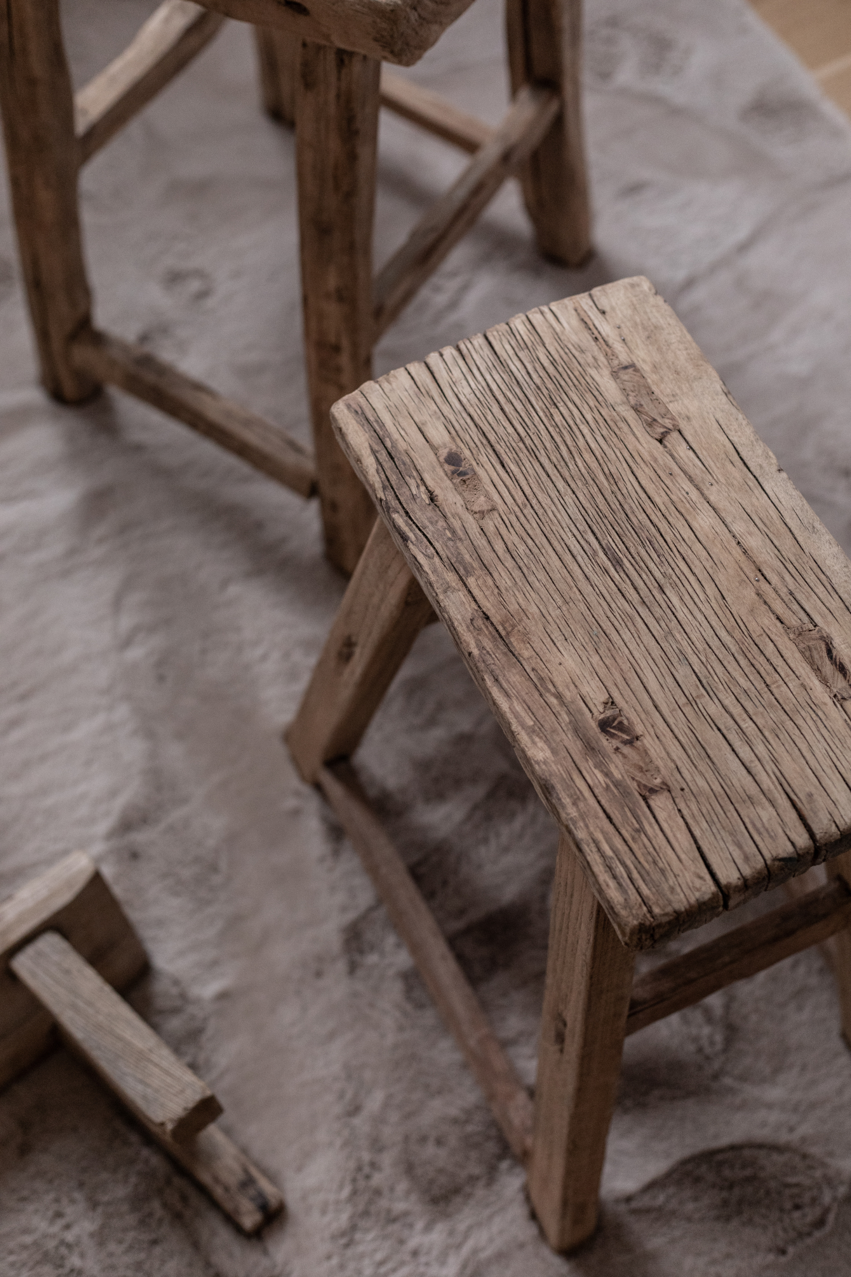 En rustik rektangulær træskammel står på et lyst tæppe. Skamlen har tydelige træårer og slidmærker, og andre små skamler ses delvist i baggrunden.