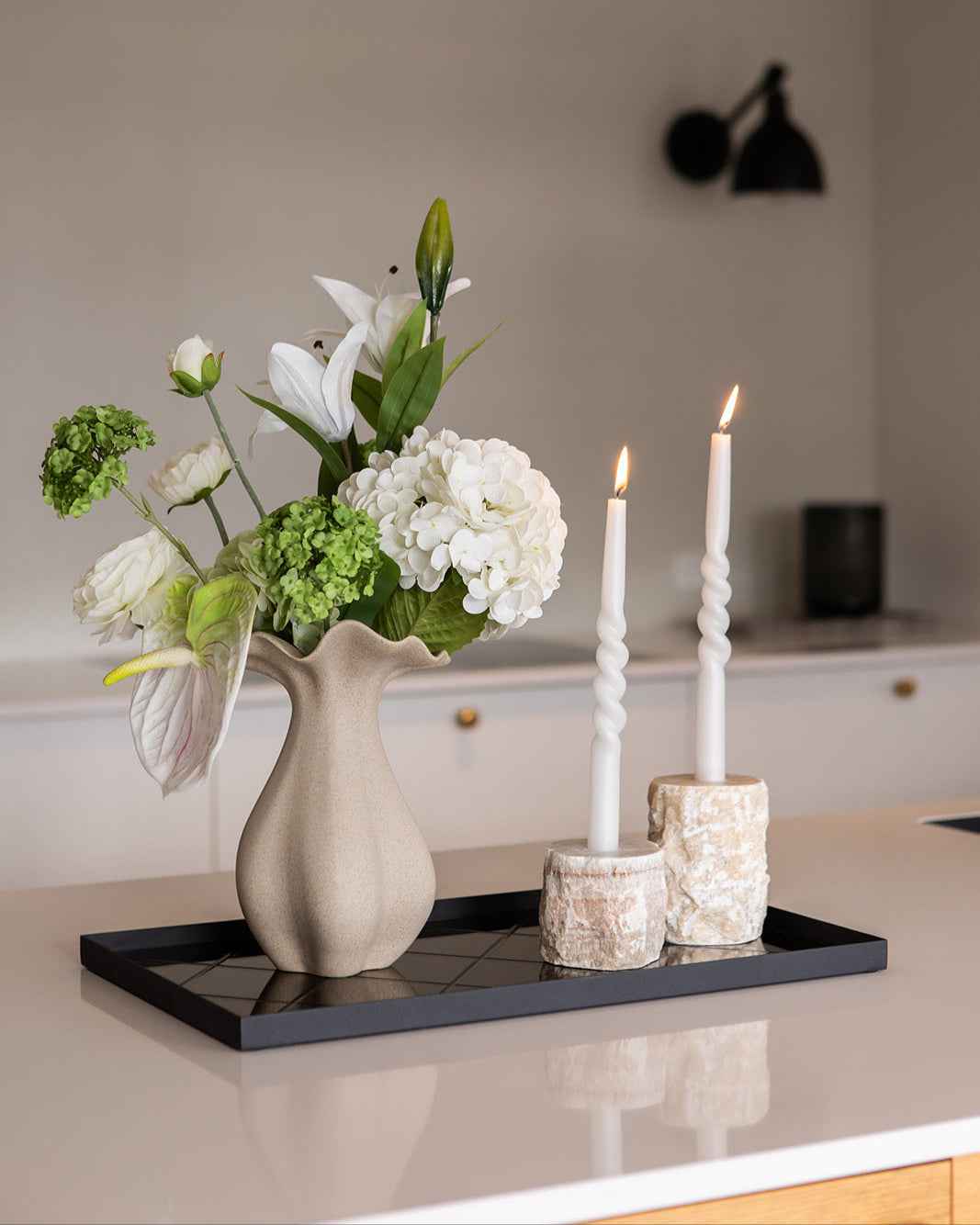 Sandfarvet keramikvase med en grøn og hvid kunstig buket i. Vasen er placeret på en spejlbakke sammen med 2 lysestager lavet af naturstenen onyx.