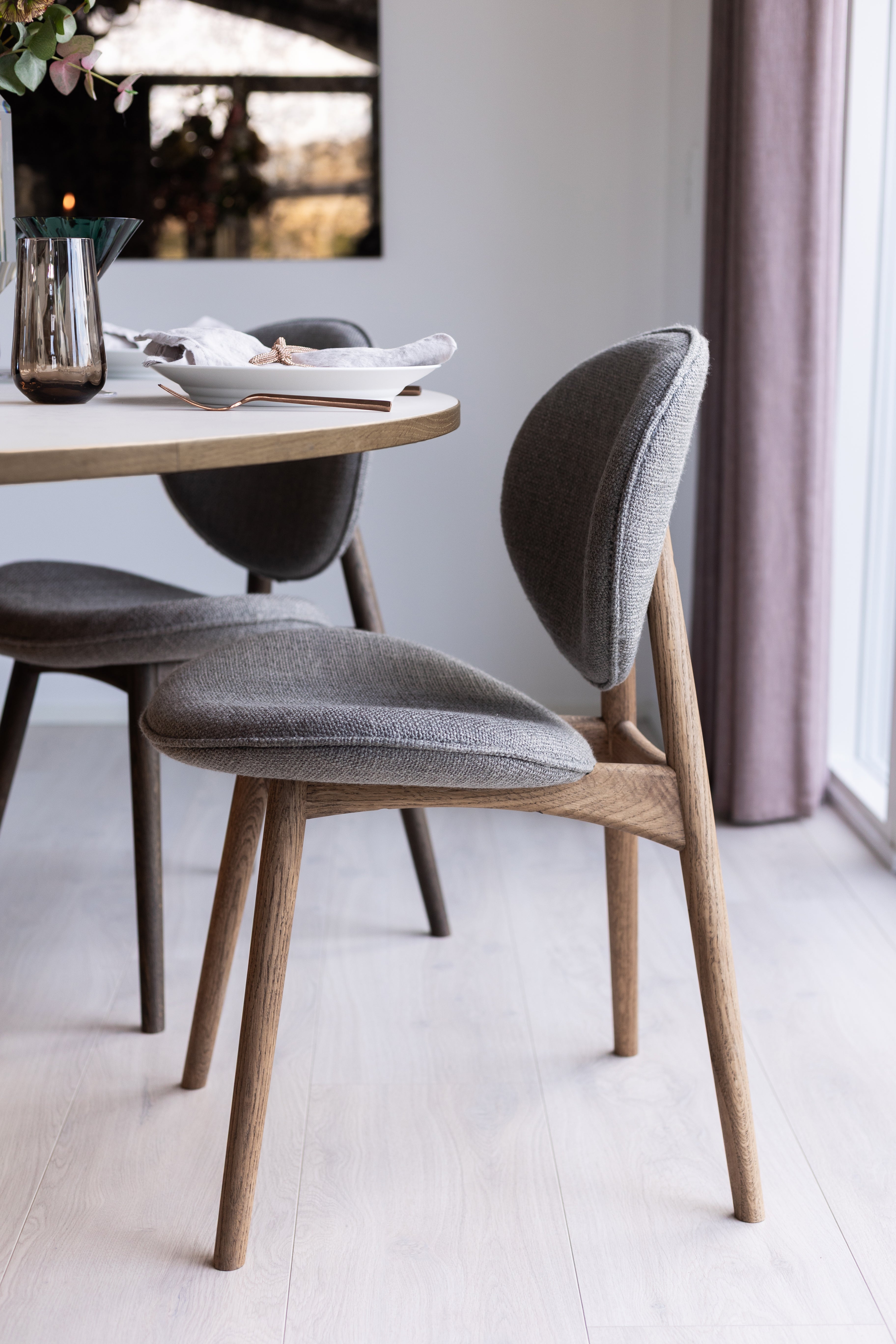 Nohr spisebordsstol i brunolieret eg med grå tekstil omkring et spisebord stylet med andre produkter fra Specktrum.