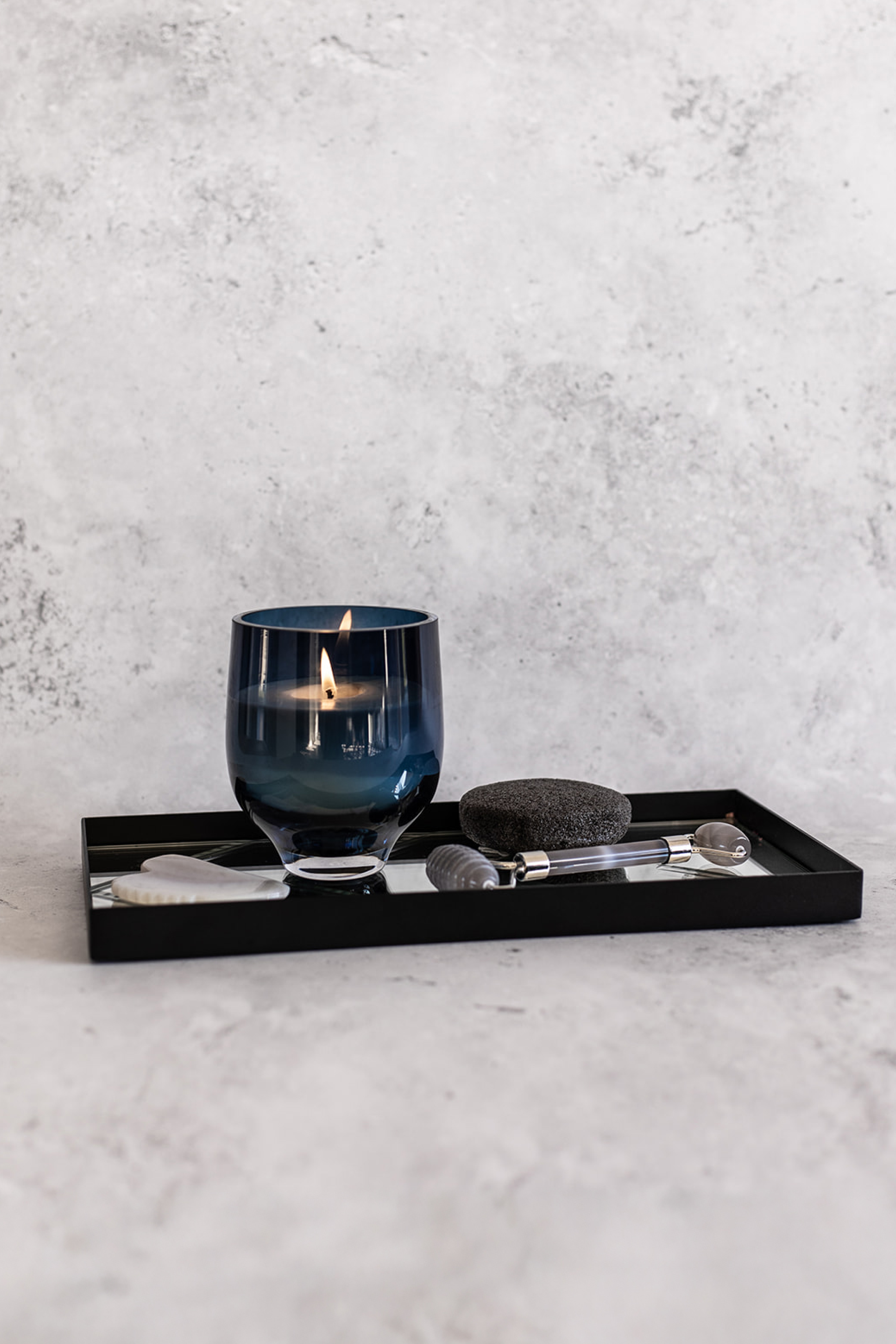 Et tændt duftlys i mørkeblåt glas står på et sort rektangulært fad med spejlbund. Ved siden af ligger en hvid gua sha-sten, en sort pumice-sten og en grå ansigtsrulle.
