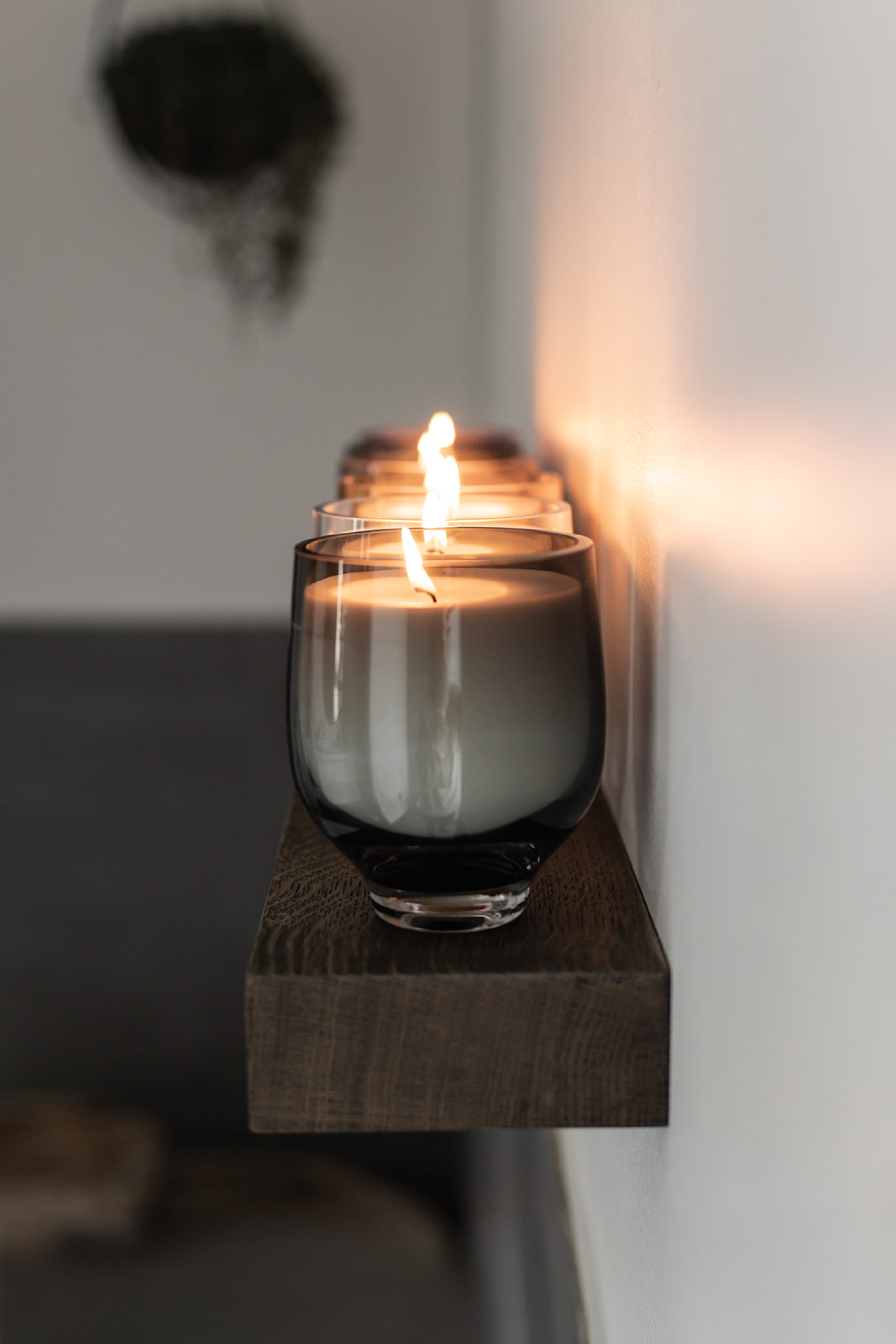 En række tændte duftlys i grå glasbeholdere står på en smal mørk træhylde langs en hvid væg.