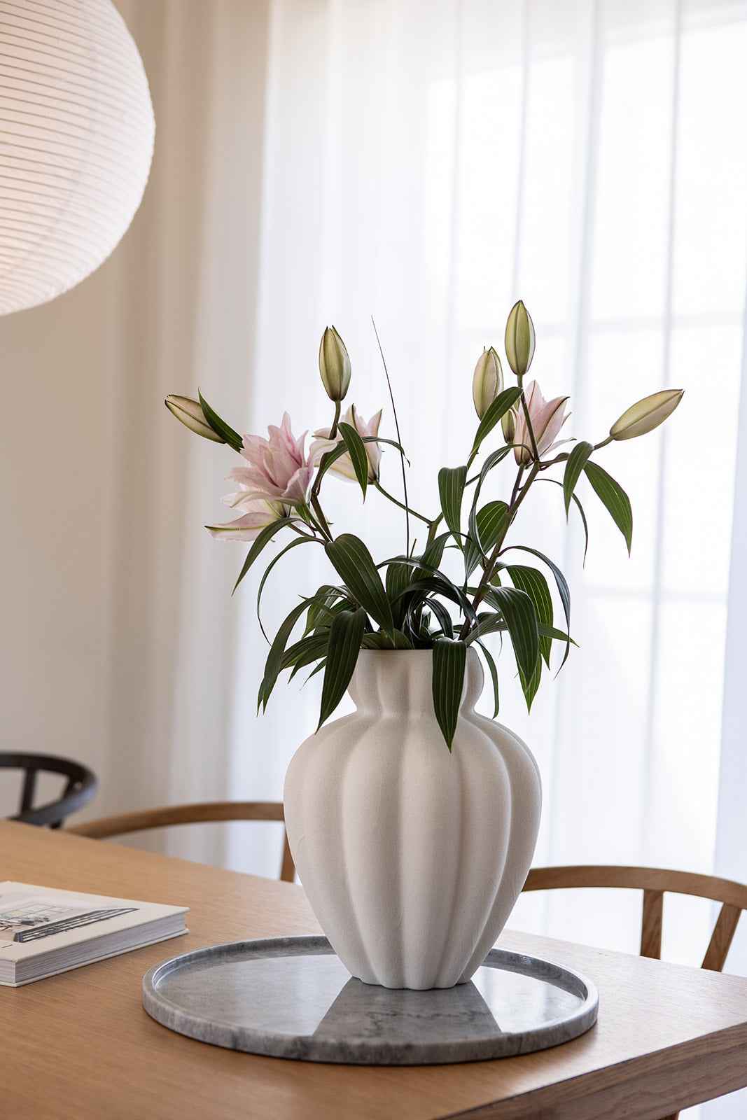 Træbord med en rund bakke i grå marmor, dekoreret med en hvid vase med lyserøde blomster og grønne blade.