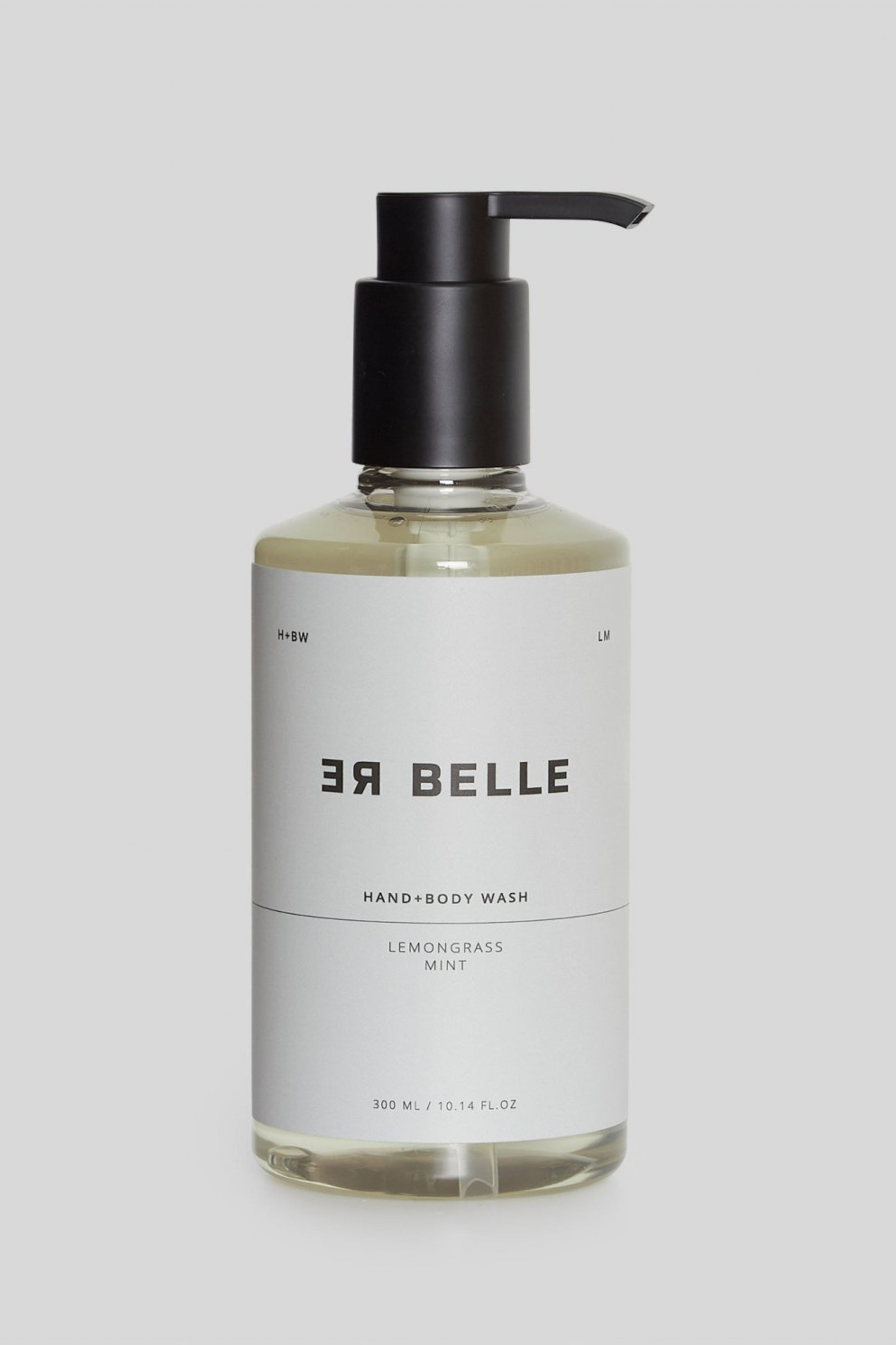 En gennemsigtig plastflaske med sort pumpe indeholdende en klar væske. Etiket i hvidt med sort tekst: “ER BELLE HAND+BODY WASH, LEMONGRASS MINT, 300 ML