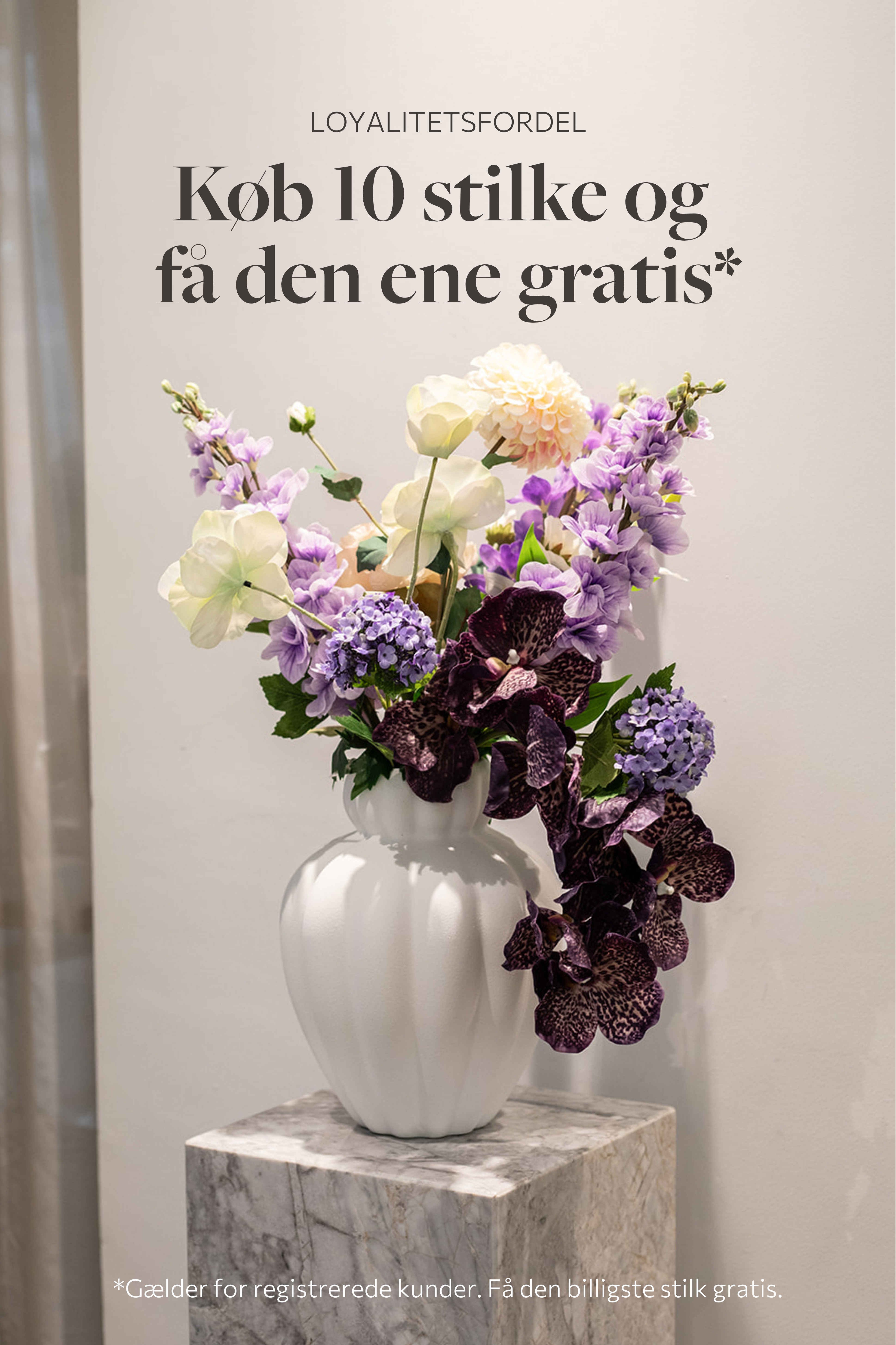 Stor buket af kunstige blomster i lilla og hvide nuancer. Placeret i en stor hvid keramikvase, der står på en marmorpedestal.