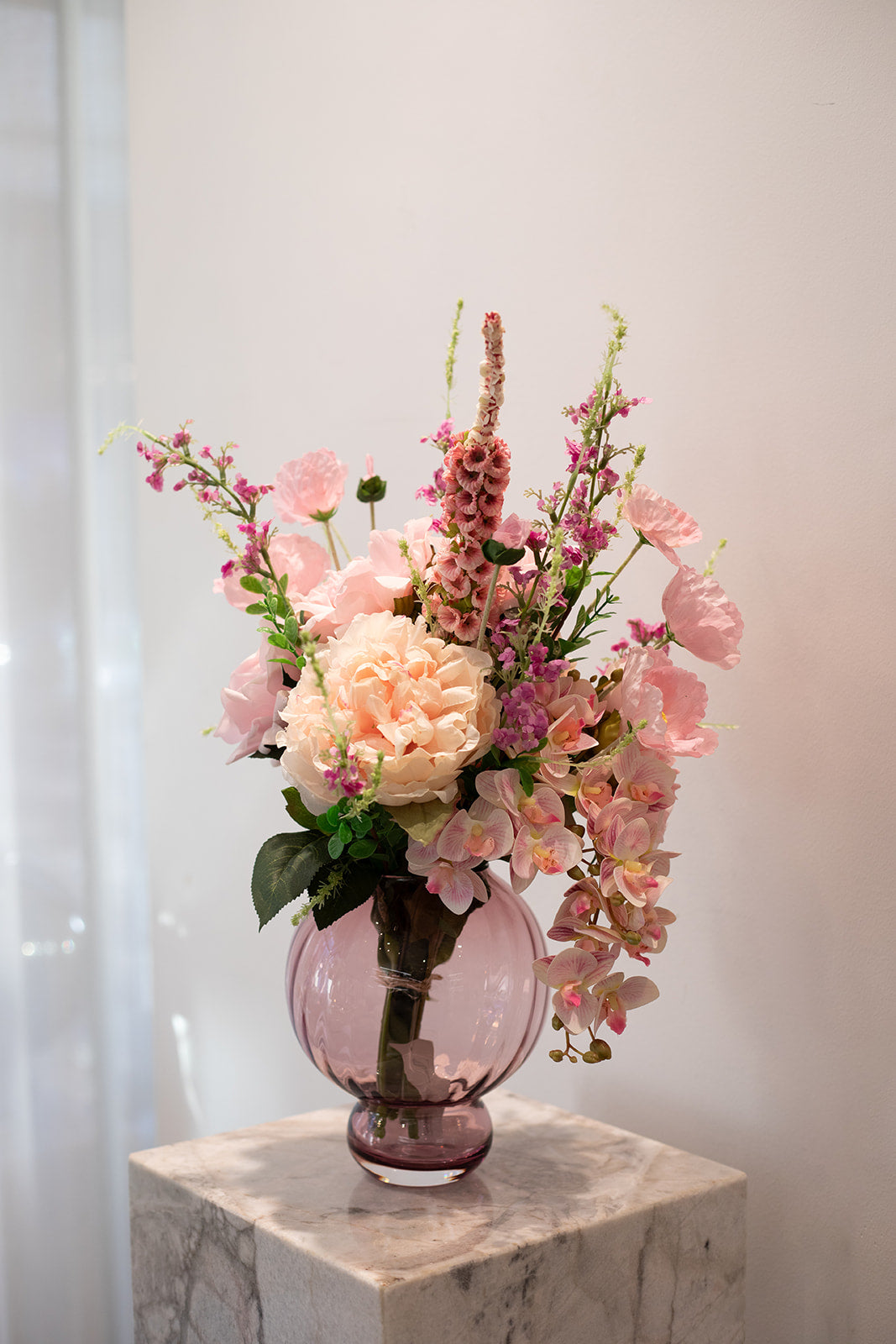 Kunstig buket i lyserøde og creme nuancer. Placeret i en lyserød glasvase, der står på en marmor pedestal.