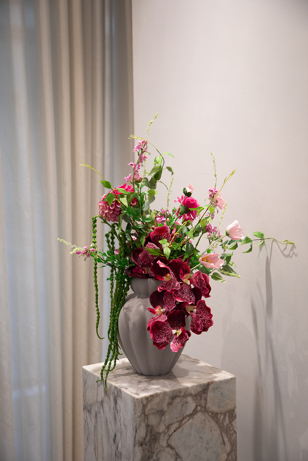 Billede af en buket i rosa og grønne nuancer af kunstige blomster. Placeret i en keramikvase, der står på en marmor pedestal.