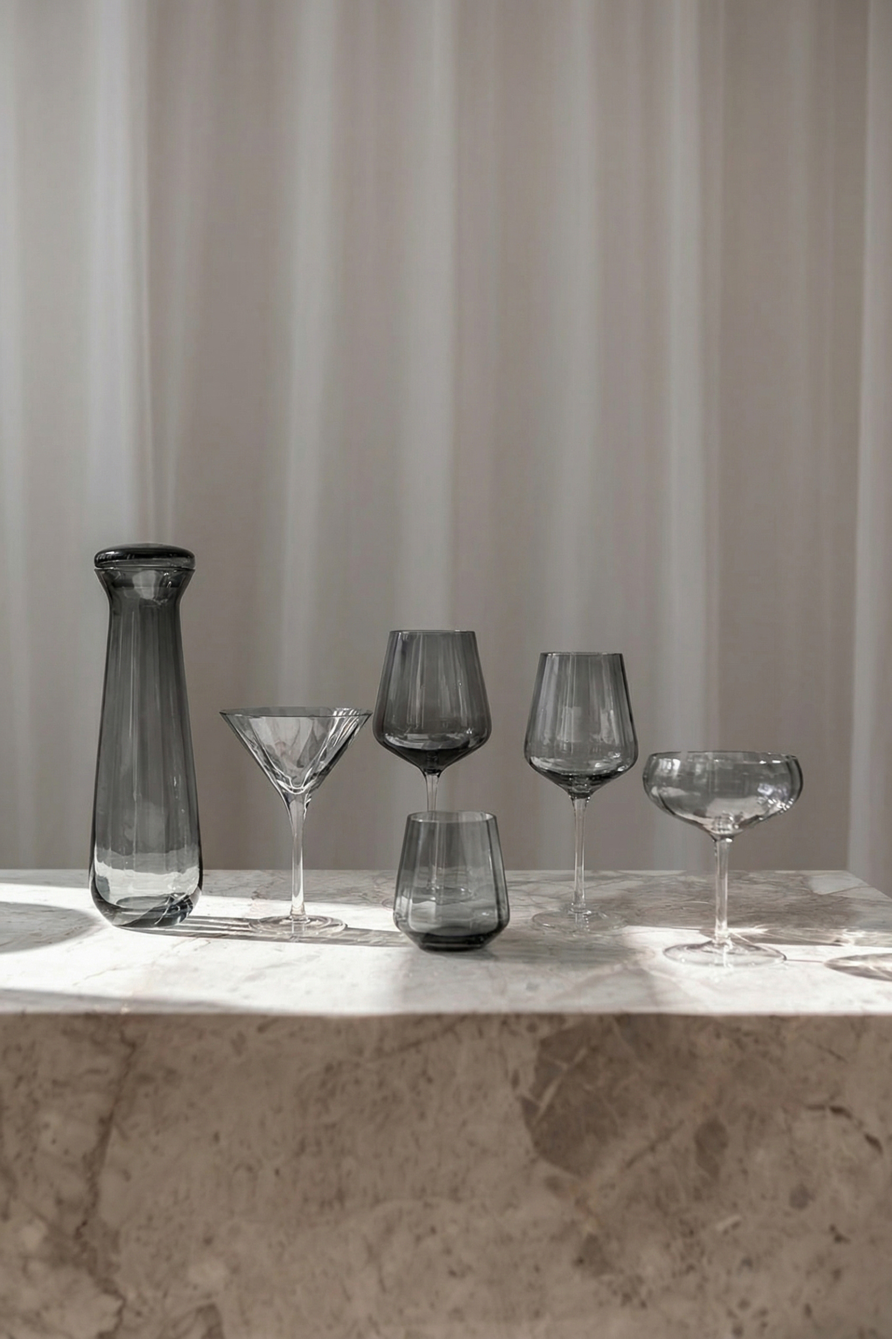 Rødvin og hvidvinsglas, cocktail glas, martini glas, drikkeglas og karaffel alle i grå farve, der står på et marmorbord.