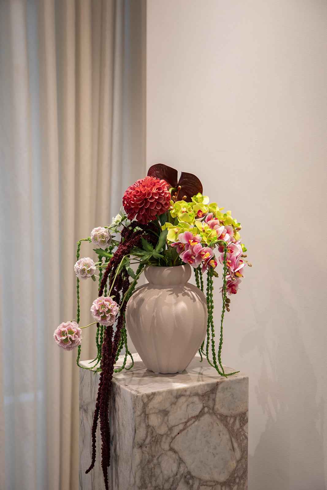 Stor farverig buket af kunstige blomster i røde, grønne og lyserøde nuancer. Placeret i en sandfarvet keramikvase, der står oven på en marmor pedestal.