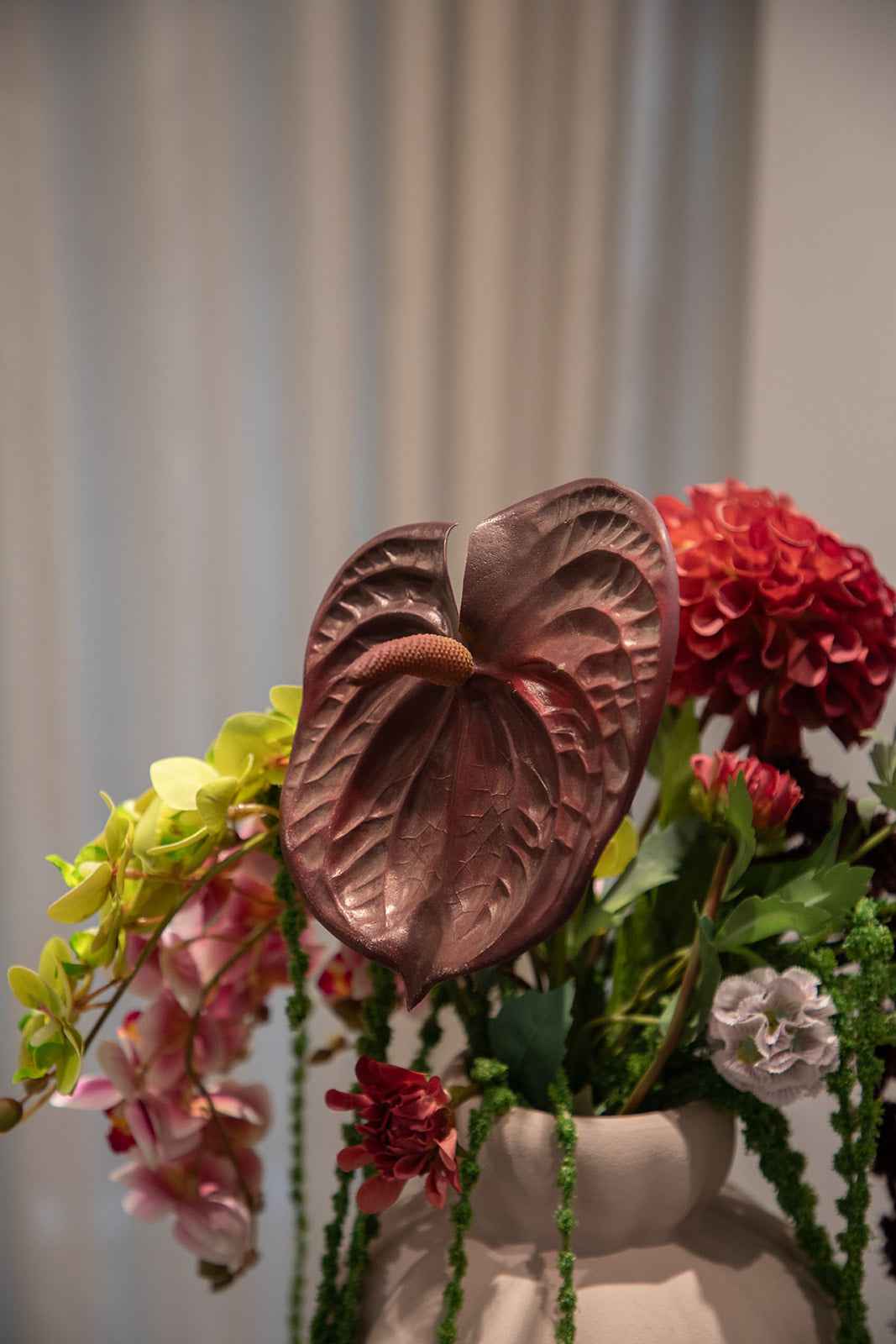 Nærbillede af en farverig buket af kunstige blomster i røde, grønne og rosa nuancer. Placeret i en sand farvede keramikvase.