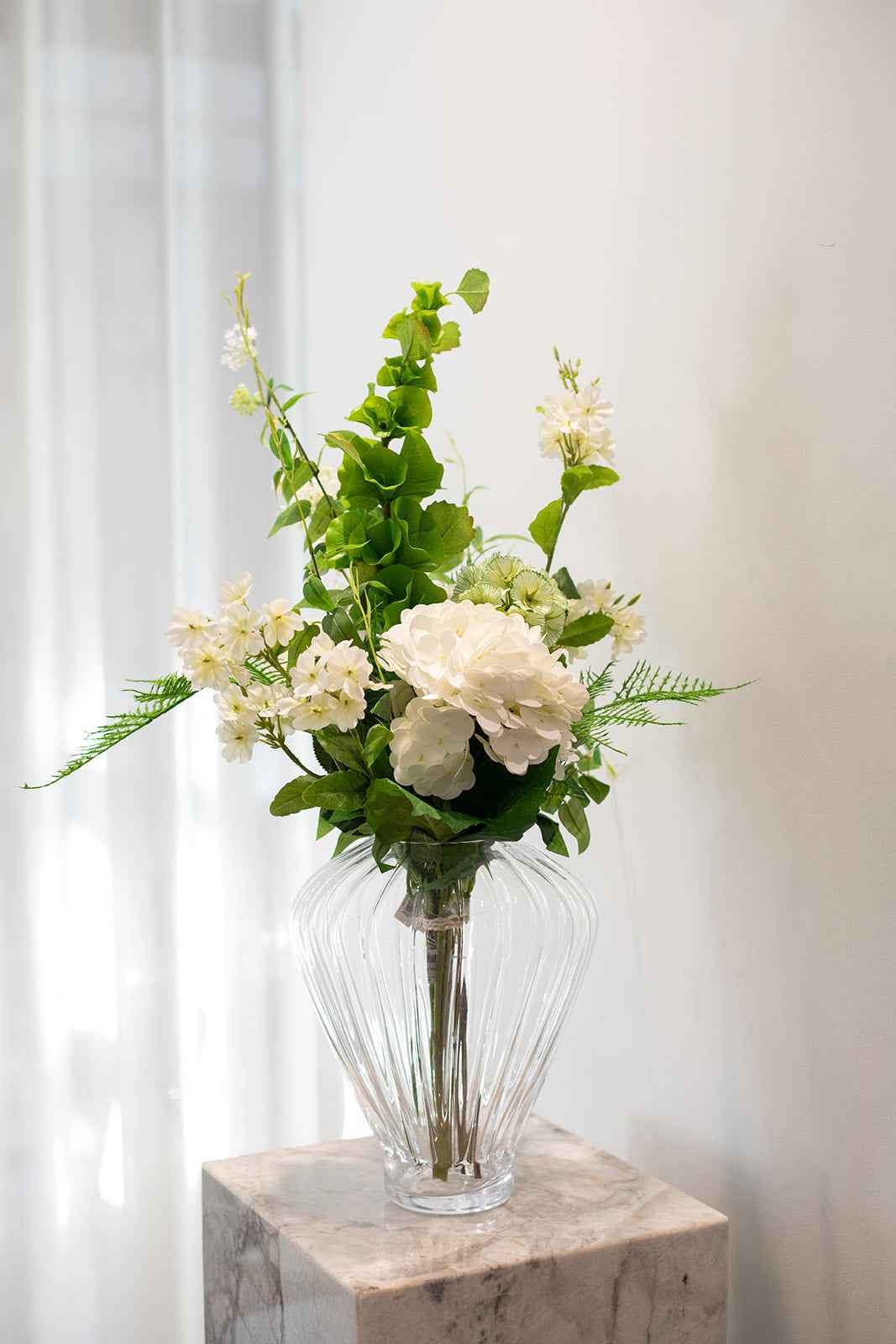 Kunstig buket i grønne og hvide nuancer, placeret i en gennemsigtig glas vase, der står på en marmor pedestal.