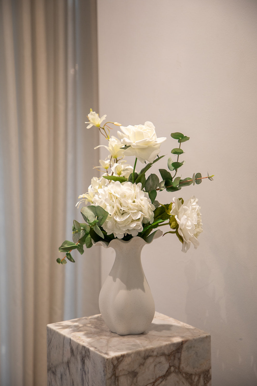 Buket af kunstige blomster i hvide og grønne nuancer. Placeret i en hvid keramik vase, der står på en marmor pedestal.